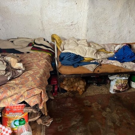 Zwei einfache Betten stehen in einer L-Form zueinander in einem Raum, an dem die Farbe von der Wand bröckelt. Die Matratzen und Bettwäsche sieht alt und schmutzig aus. Essensvorräte sind unter den Betten gelagert.