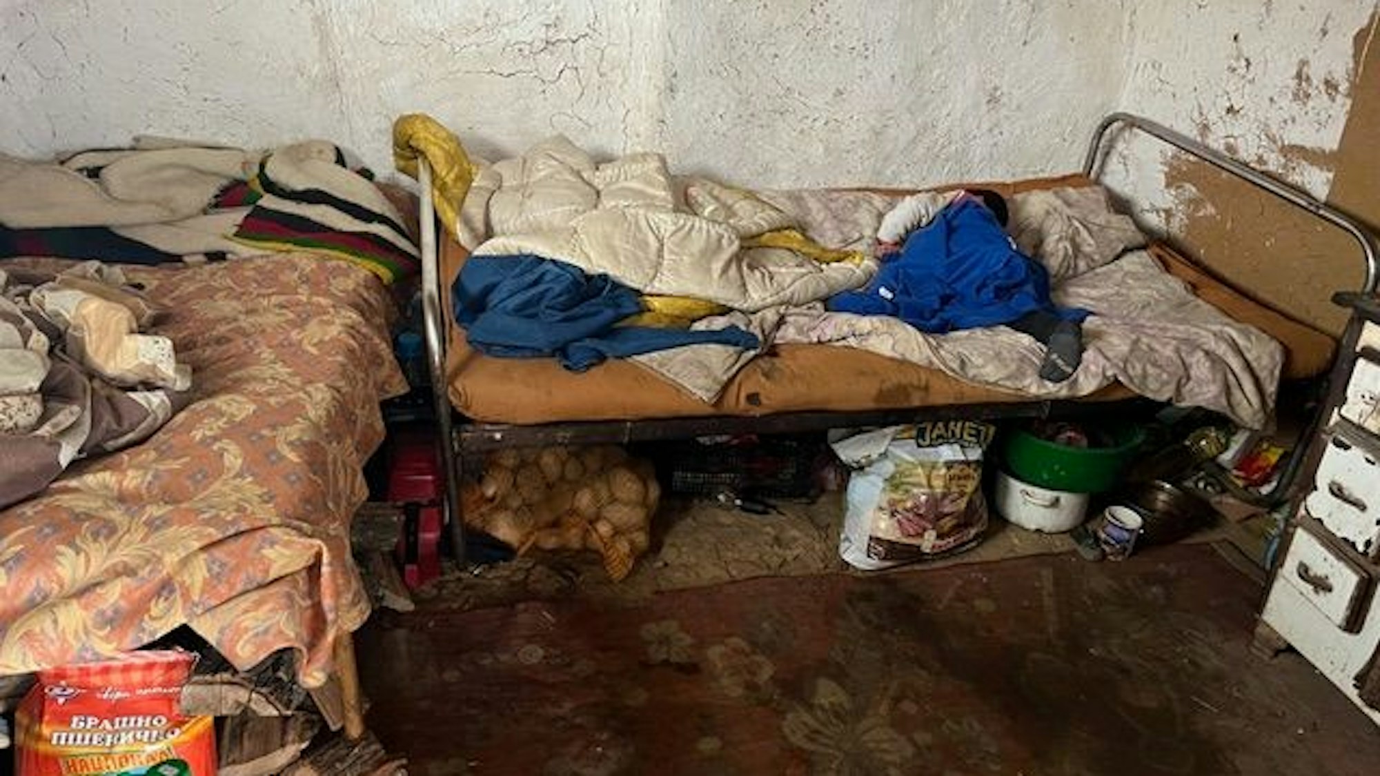 Zwei einfache Betten stehen in einer L-Form zueinander in einem Raum, an dem die Farbe von der Wand bröckelt. Die Matratzen und Bettwäsche sieht alt und schmutzig aus. Essensvorräte sind unter den Betten gelagert.