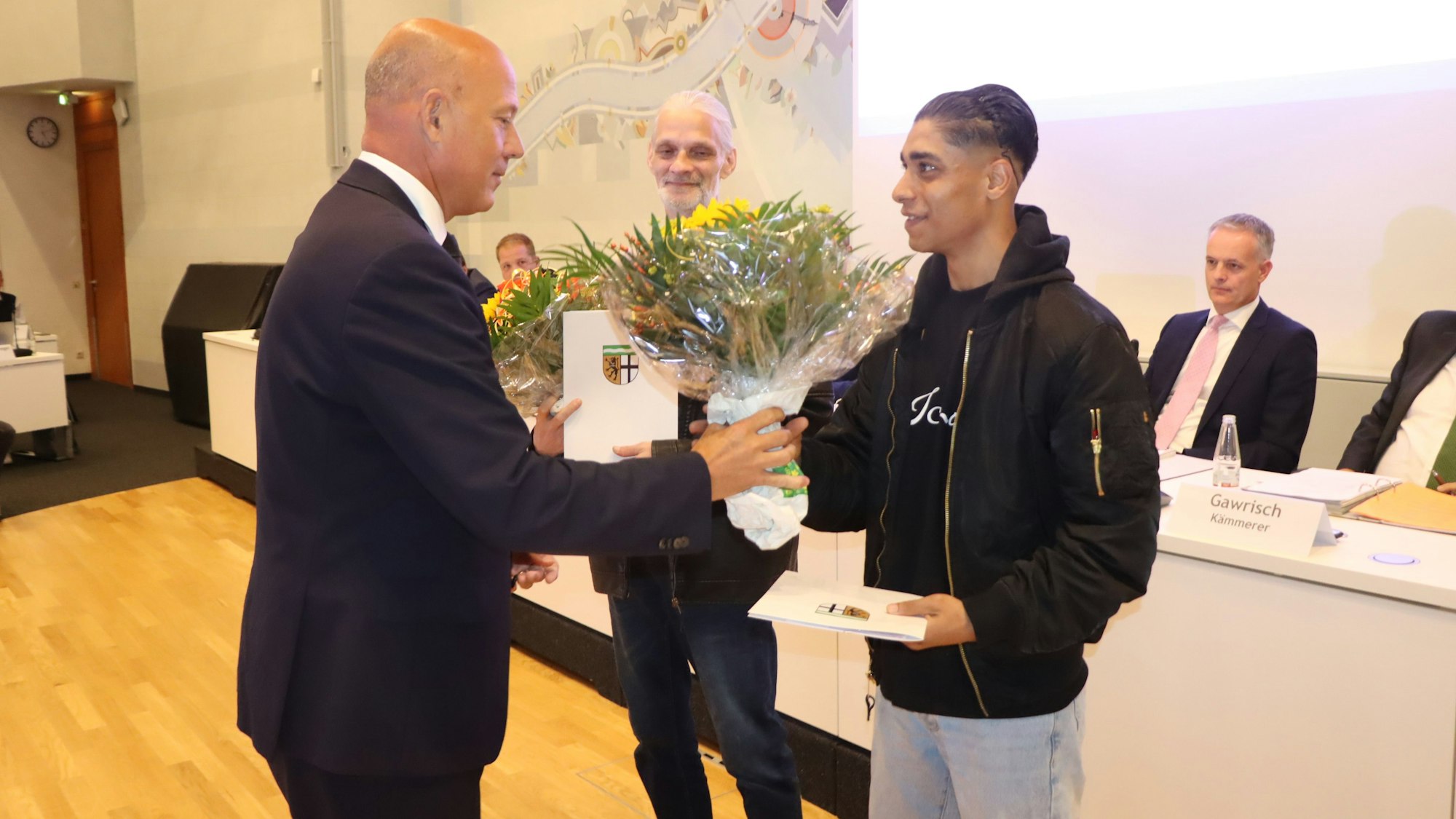 Zu sehen ist ein Mann, der einem anderen einen Blumenstrauß überreicht.