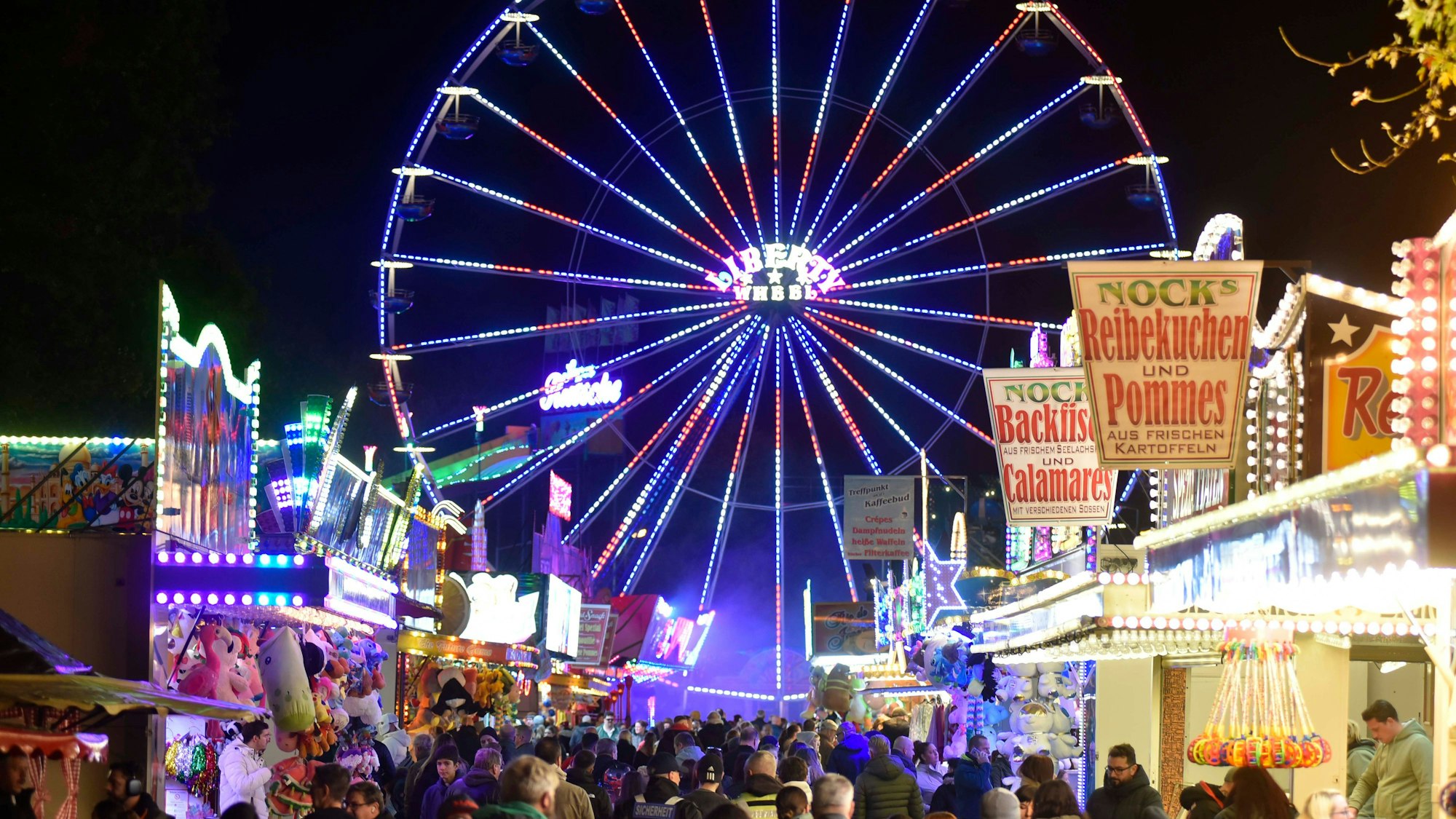 Zu sehen ist ein Riesenrad mit Abendbeleuchtung auf einem Jahrmarkt.