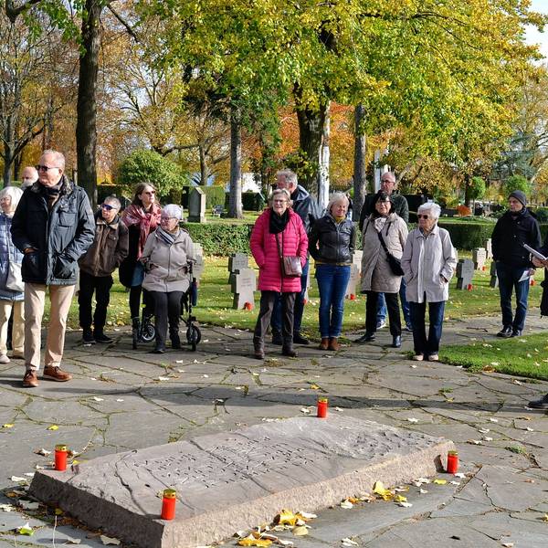 Eine Besuchergruppe steht vor einem Grab, dessen waagerechte Steintafel mit vier an den Ecken aufgestellten Kerzen geschmückt ist. Kurt Lingscheidt liest aus einem Buch vor.