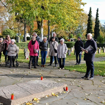 Eine Besuchergruppe steht vor einem Grab, dessen waagerechte Steintafel mit vier an den Ecken aufgestellten Kerzen geschmückt ist. Kurt Lingscheidt liest aus einem Buch vor.
