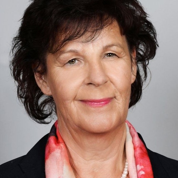 Eine Frau mit dunklem Haar, dunklem Blazer und Perlenkette schaut lächelnd in die Kamera.