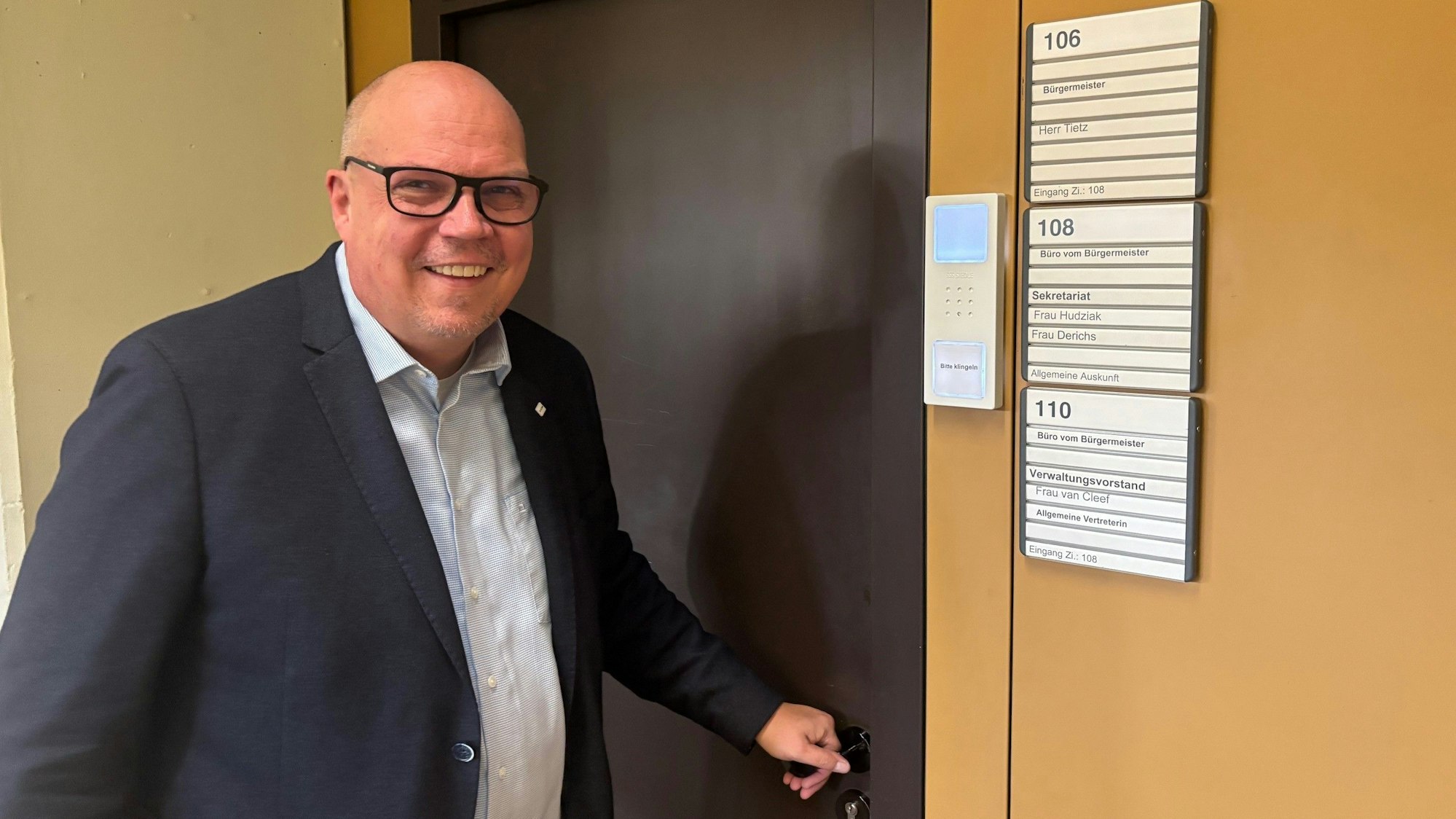 Auf dem Bild ist ein Mann im blauen Sakko zu sehen, der eine braune Bürotür öffnet.