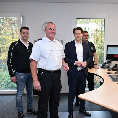 Vier Menschen stehen am Schaltpult in einer Feuerwehr-Leitstelle.