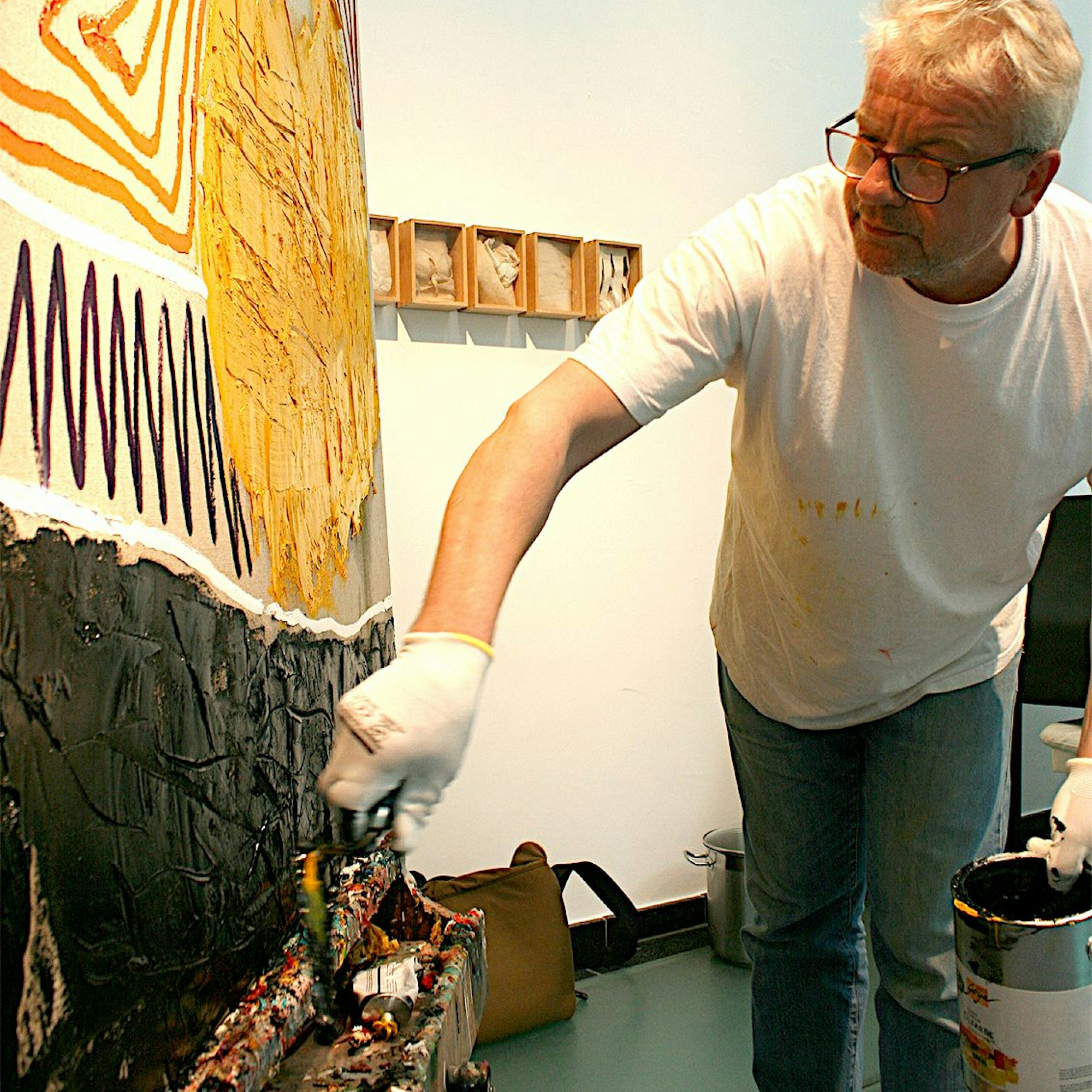 Der Künstler steht an einer Leinwand, in der Hand hält er einen Eimer Farbe, mit der anderen spachtelt er eine Schicht schwarze Farbe auf.