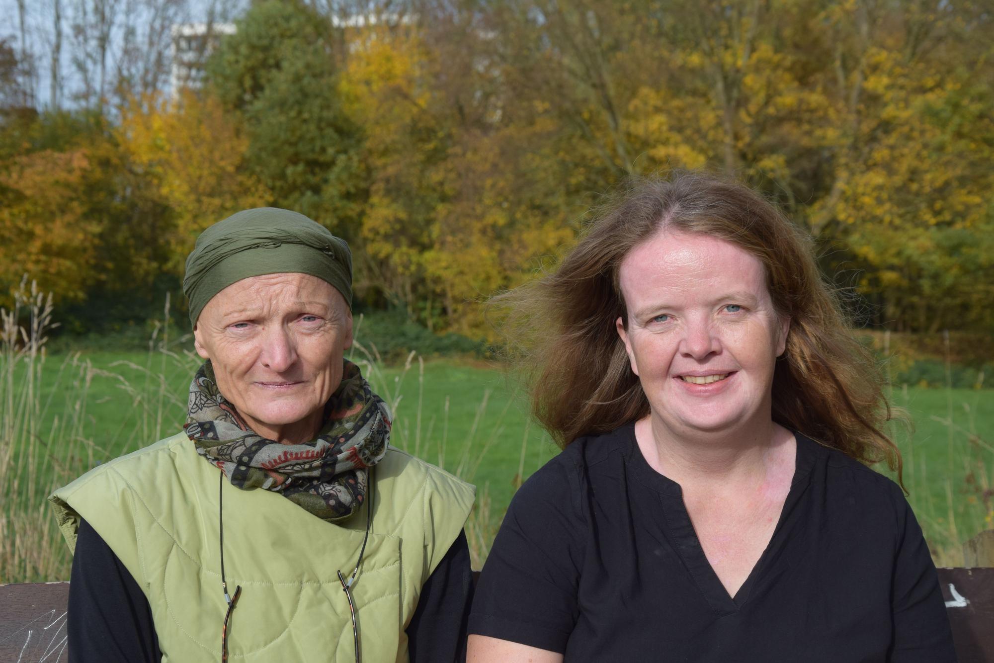 Eine Frau mit grünem Hut und eine jüngere Frau mit langem braunem Haar sitzen nebeneinander auf einer Bank.