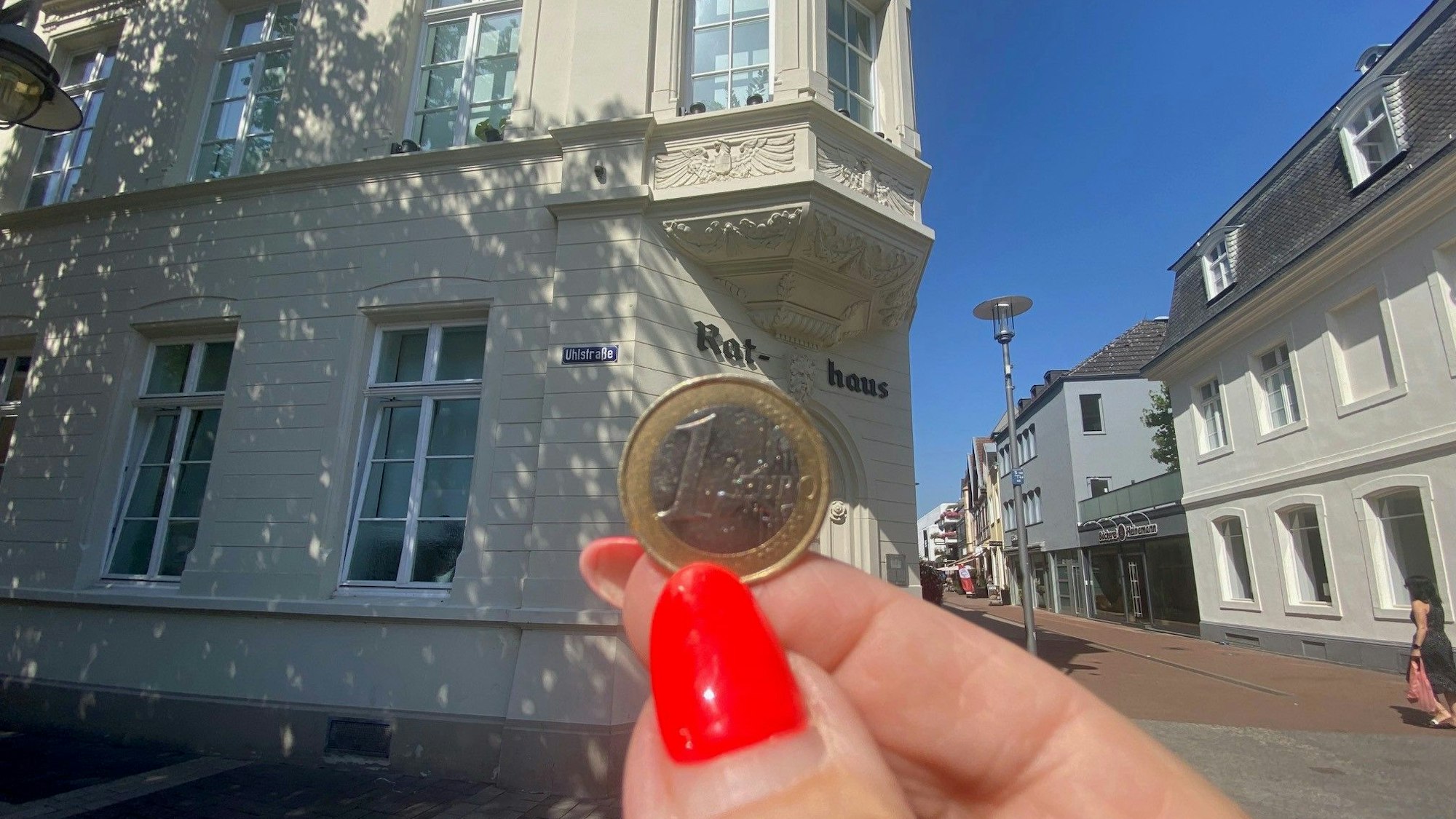 Zu sehen ist eine Hand, die eine Münze hält. Im Hintergrund steht das Brühler Rathaus.