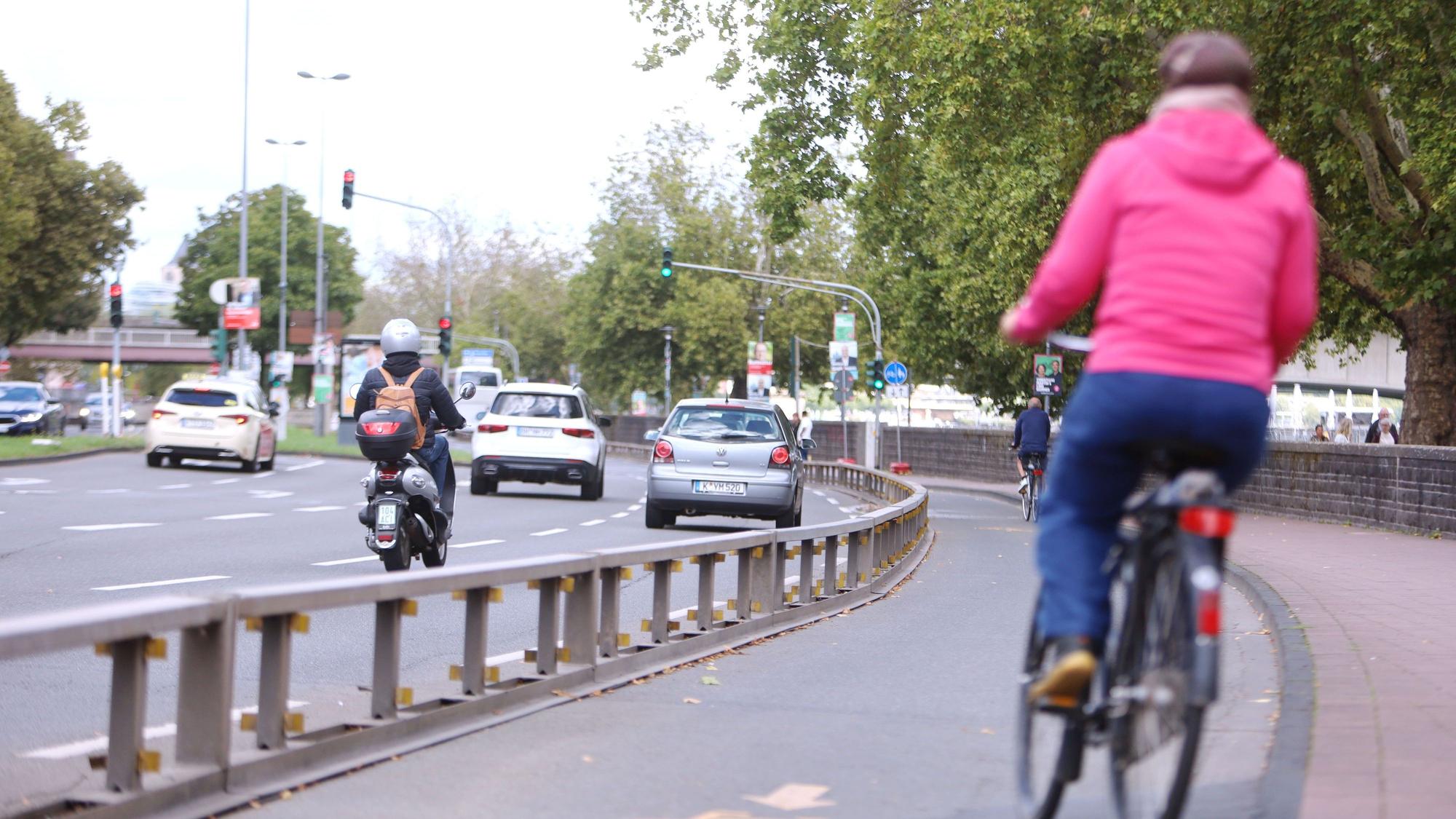 Radfahrerin fährt im Vordergrund auf dem provisorischen Radweg, der auf einer alten Fahrspur für Kfz eingerichtet ist. Abgetrennt von einer kleinen Absperrung fahren daneben Autos und ein Roller.