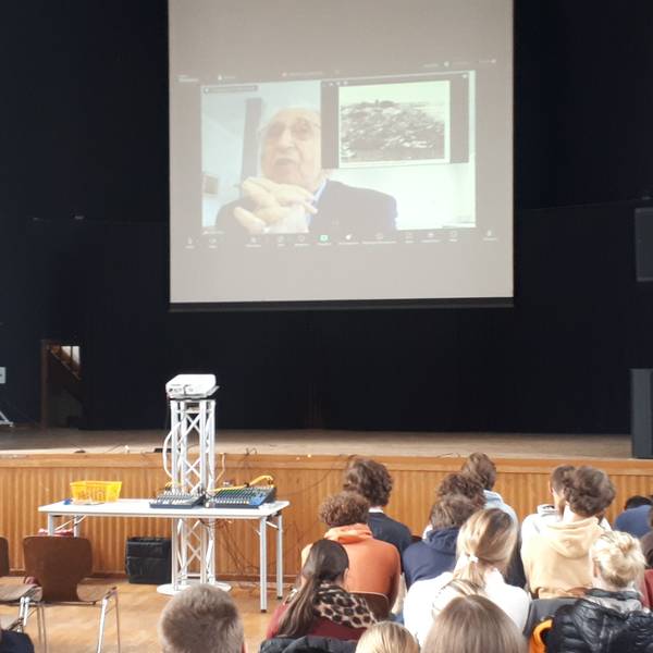 In einer Schulaula hängt über der Bühne eine Leinwand, auf der ein alter Mann zu sehen ist. Er beantwortet Fragen der Schüler, die in der Aula sitzen.