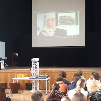 In einer Schulaula hängt über der Bühne eine Leinwand, auf der ein alter Mann zu sehen ist. Er beantwortet Fragen der Schüler, die in der Aula sitzen.