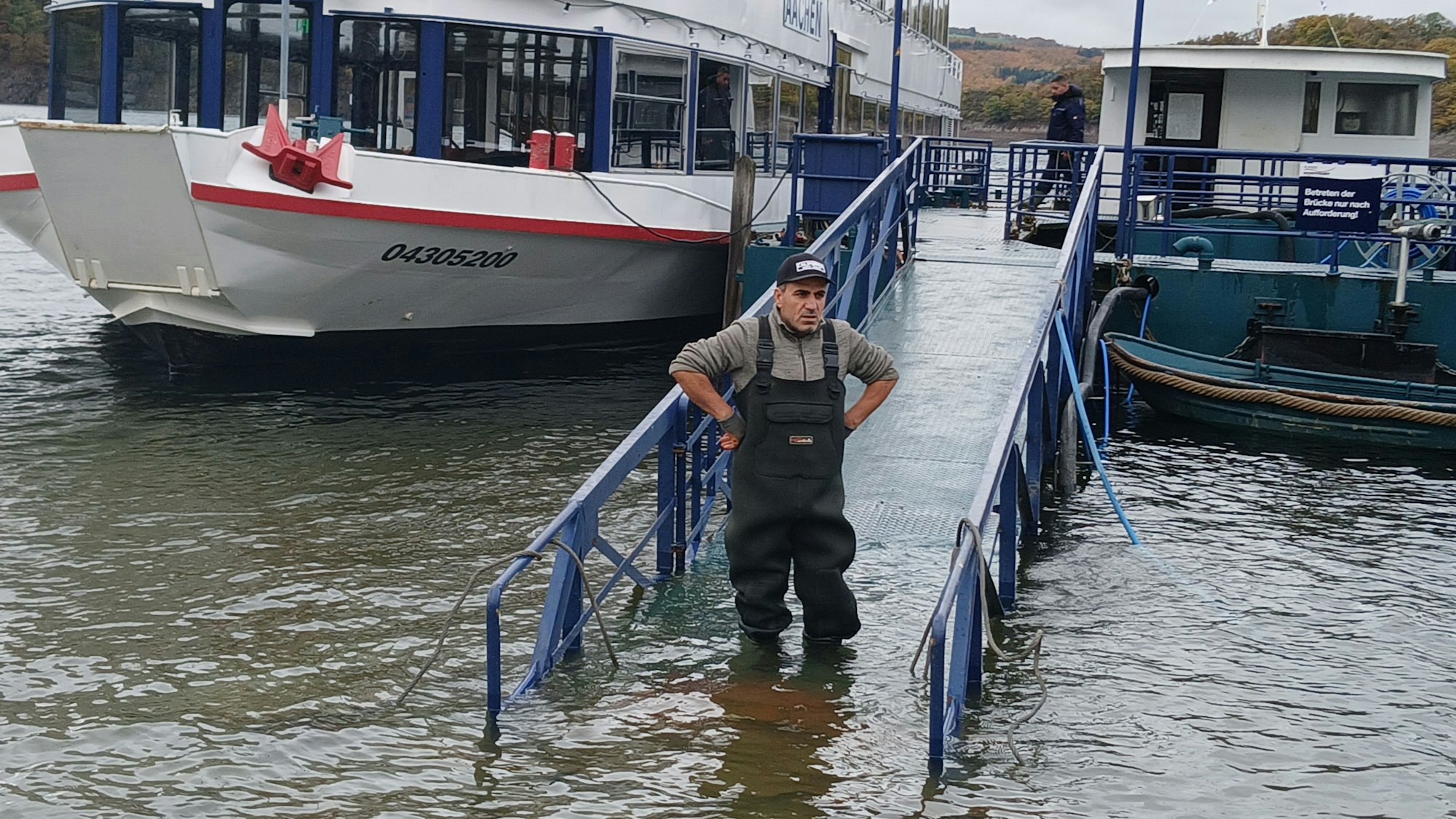 Ein Mann in Overall steht im Wasser, das Teile eines Stegs überflossen hat. Er hat die Hände in die Seiten gestemmt und schaut missmutig vor sich hin.