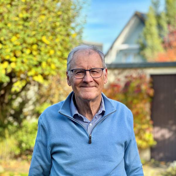 Ein Mann mit Brille, blauem Pullover und grauen Haaren steht in einem Garten.