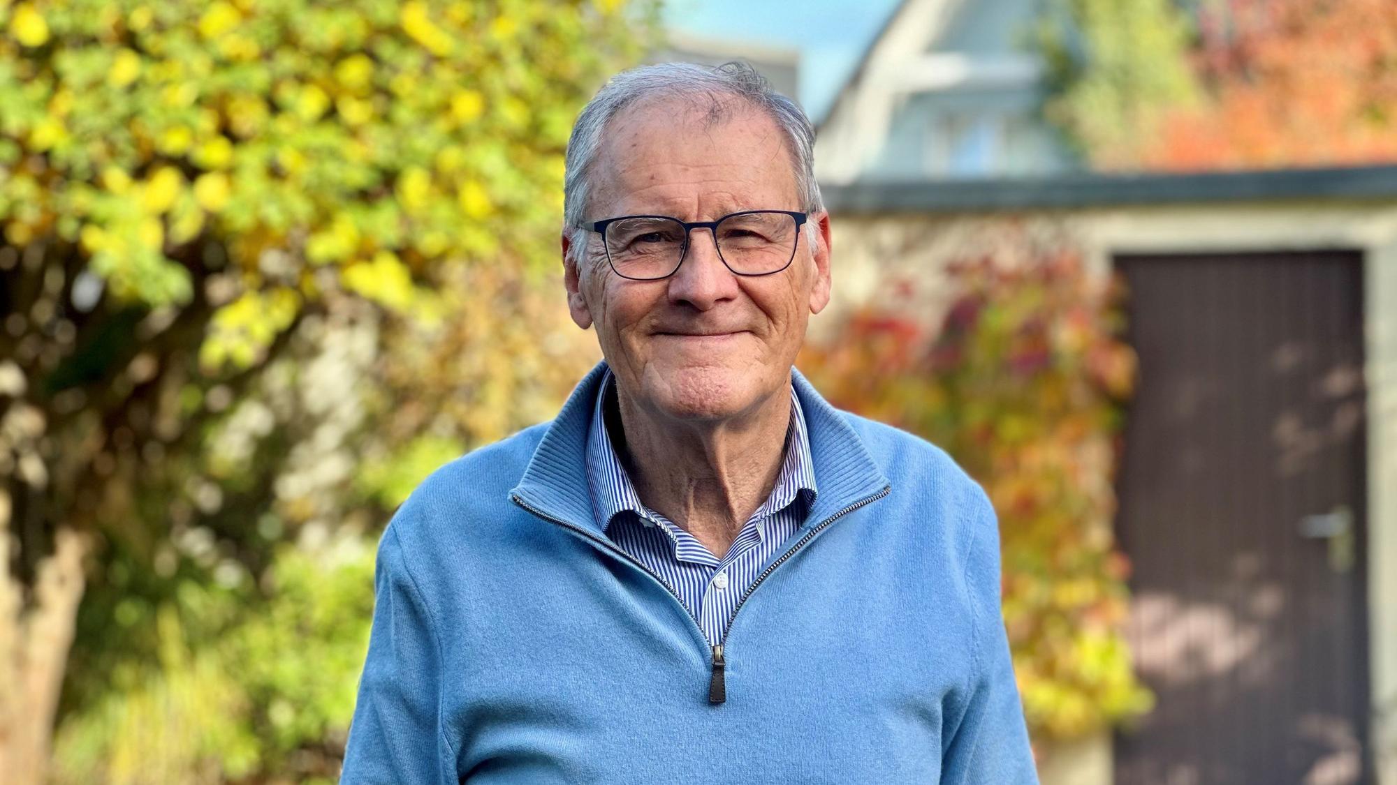 Ein Mann mit Brille, blauem Pullover und grauen Haaren steht in einem Garten.