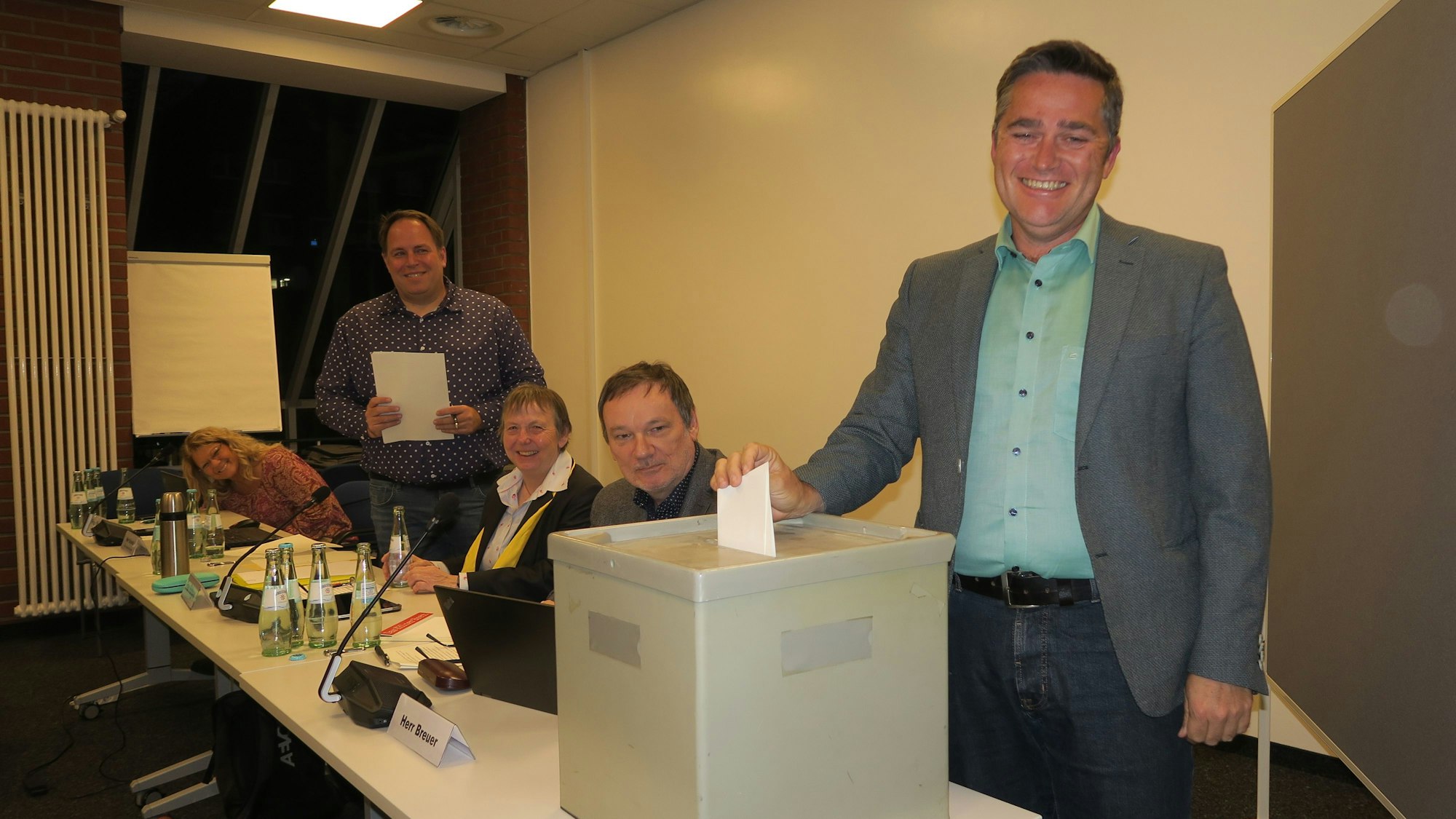 Ein Mann steht neben einer Wahlurne. An einem langen Tisch sitzen drei weitere Personen.