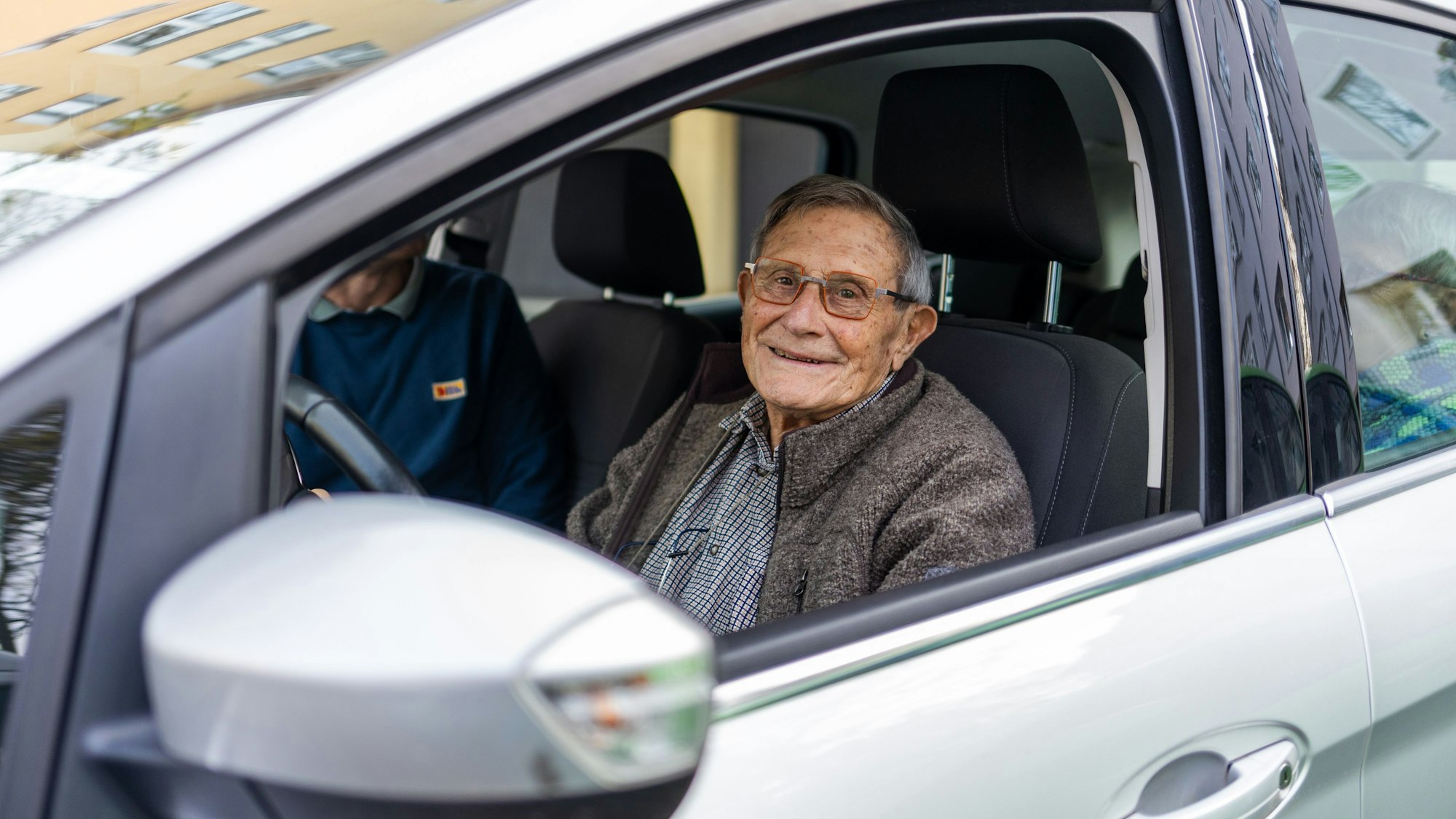 Blick durch das offene Fenster in ein Auto: Am Steuer sitzt ein älterer Mann mit Brille und brauner Jacke über dem Hemd.
