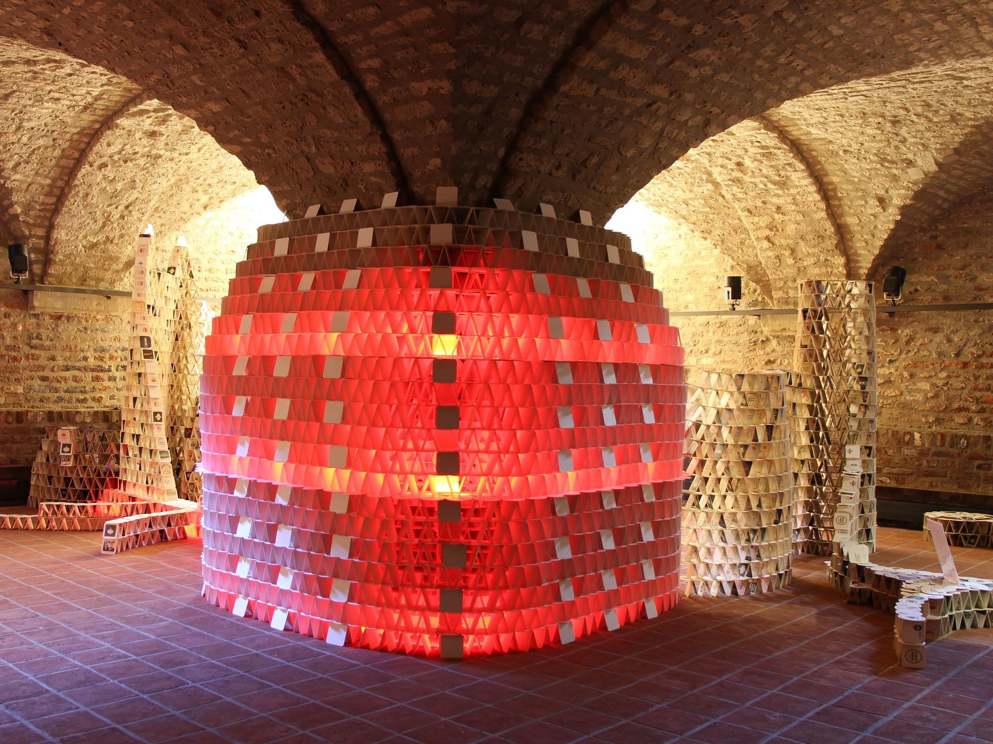 Eine mehrere Meter hohe Skulptur aus Bierdeckeln, sie ist in rotes Licht getaucht.