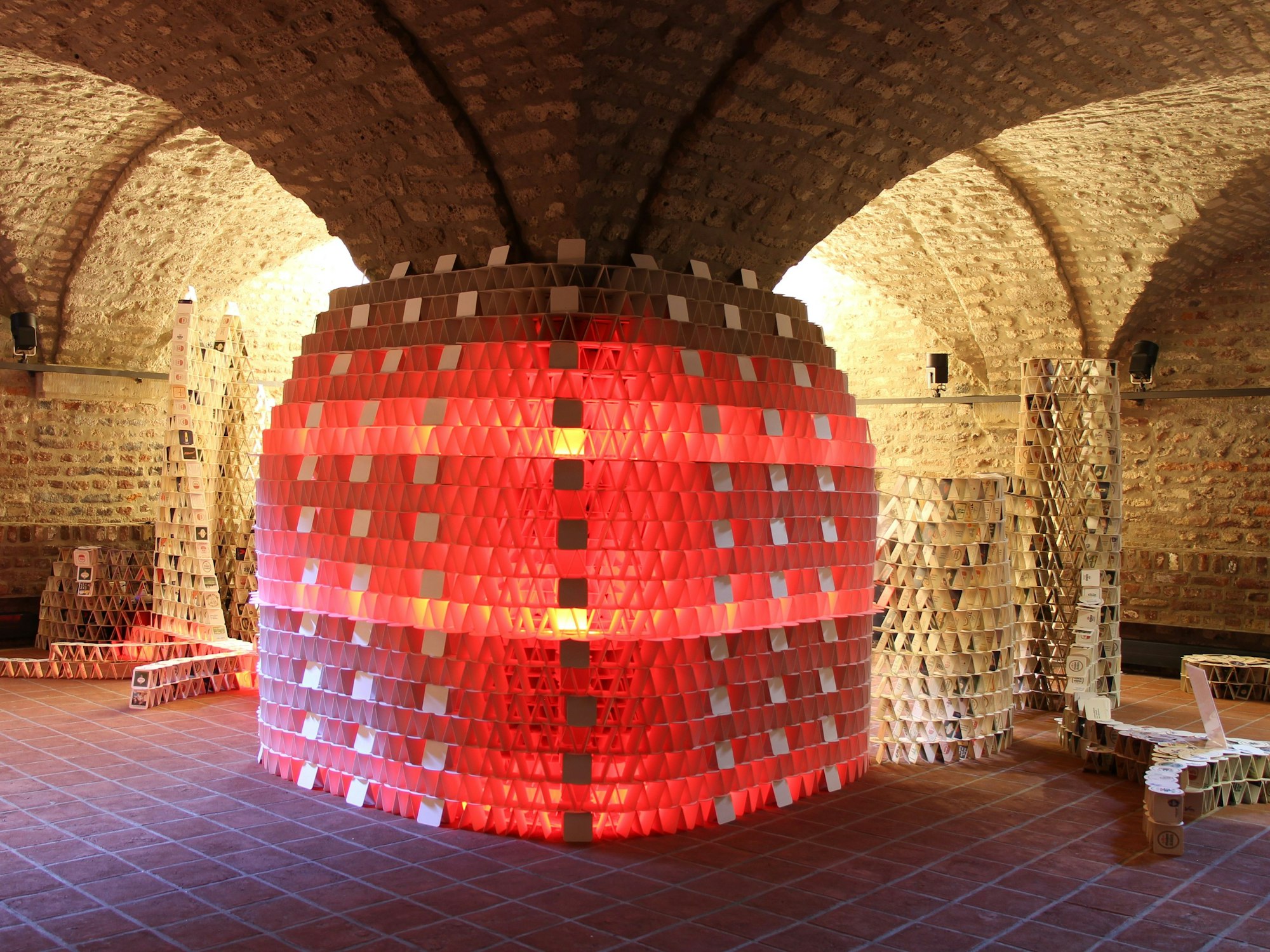 Eine mehrere Meter hohe Skulptur aus Bierdeckeln, sie ist in rotes Licht getaucht.