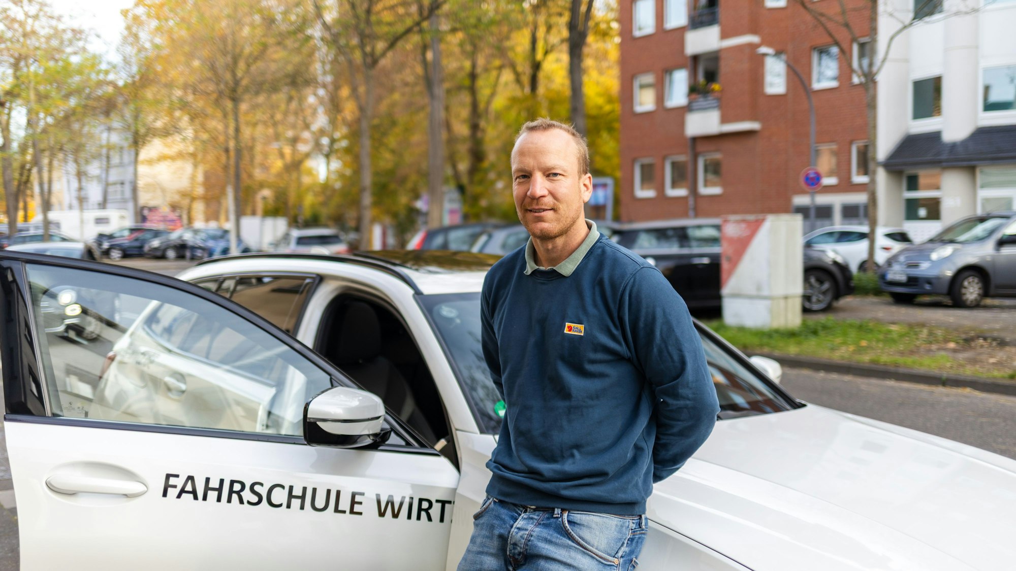 Ein Mann mit blauen Pullover lehnt an einem weißen Auto mit Aufschrift „Fahrschule Wirtz“