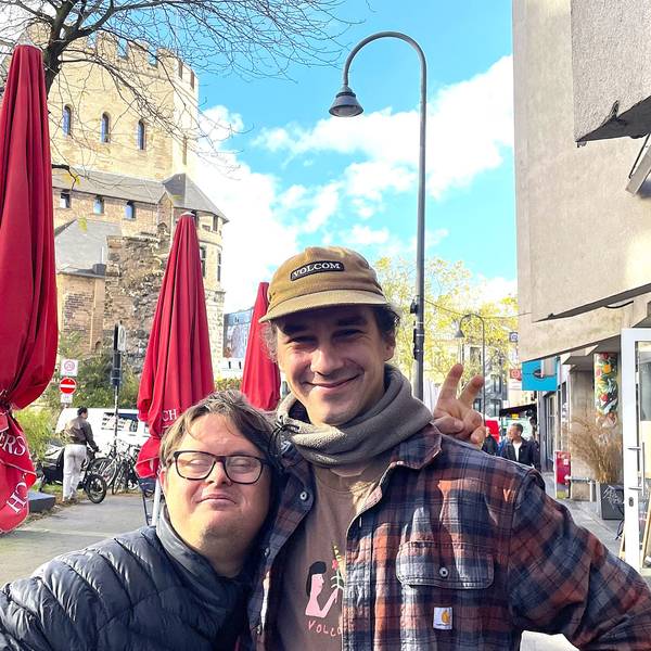 Ein kleiner Mann mit Brille wird von einem großen mit Kappe in den Arm genommen.