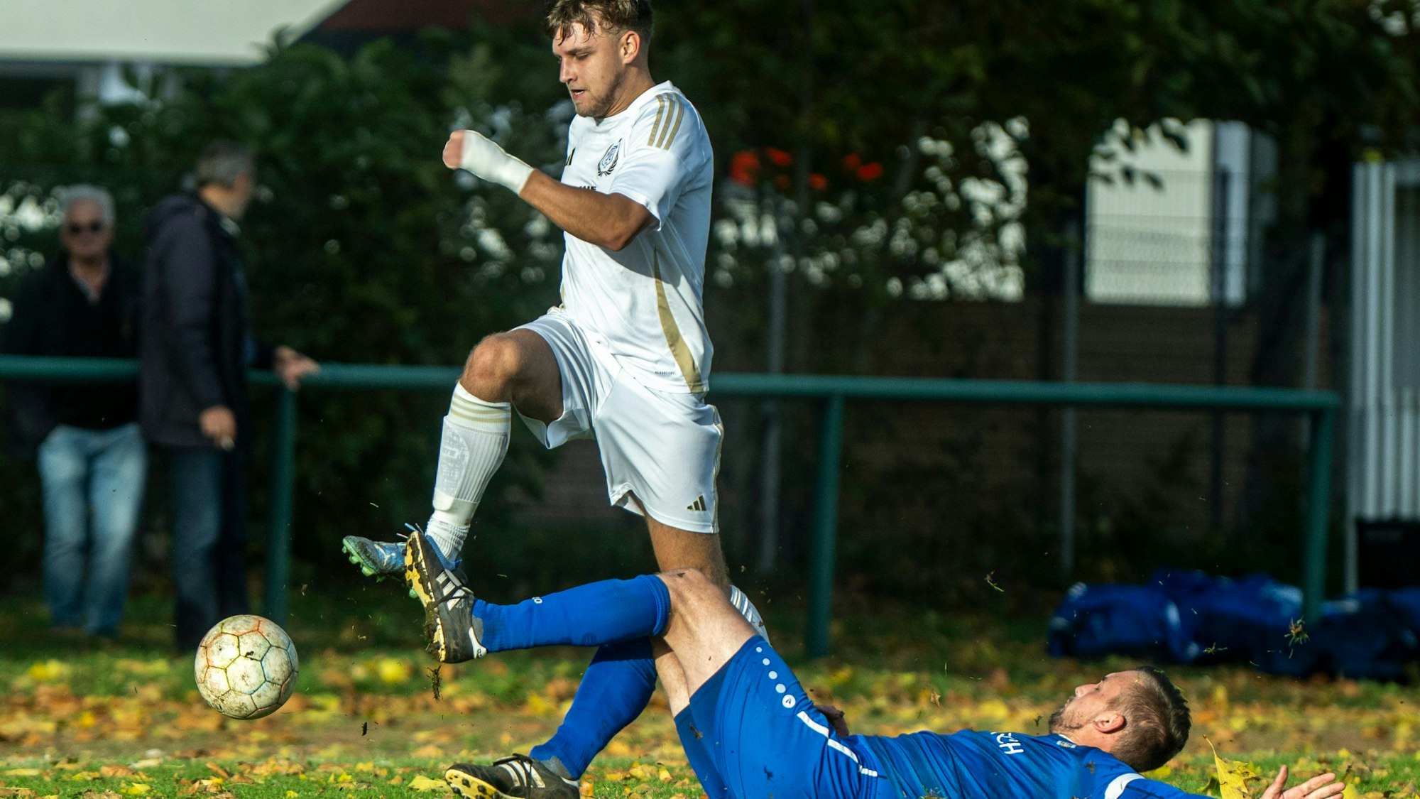 Ein Fußballspieler im blauen Triktot liegt am Boden und spielt einen Ball weg, den der Spieler im weißen Trikot so nicht mehr erreichen kann.