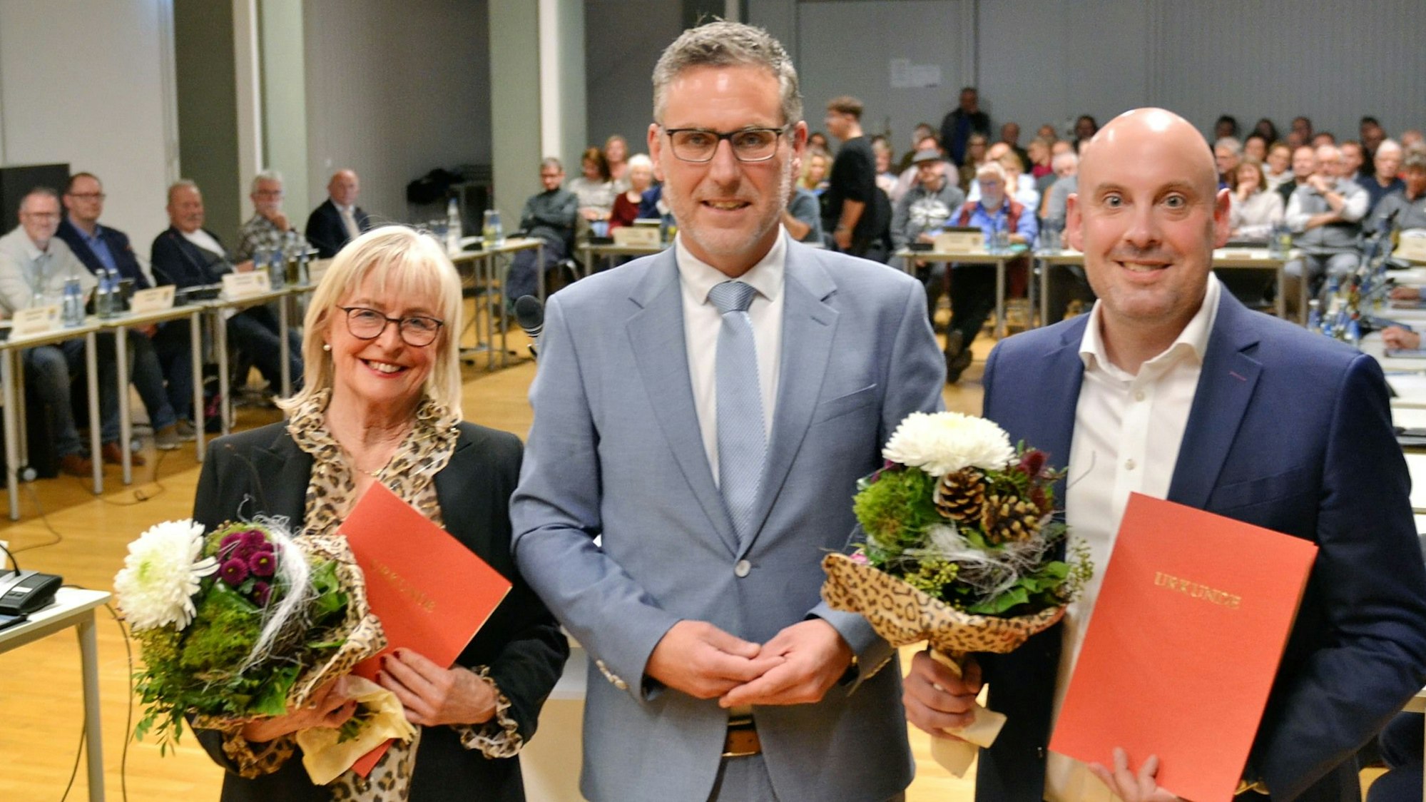 Rechts und links von Nettersheims Bürgermeister Norbert Crump stehen seine Stellvertreter. Gertrud Schruff und Andre Knips halten Blumen und rote Mappen in der Hand, auf denen – steht.