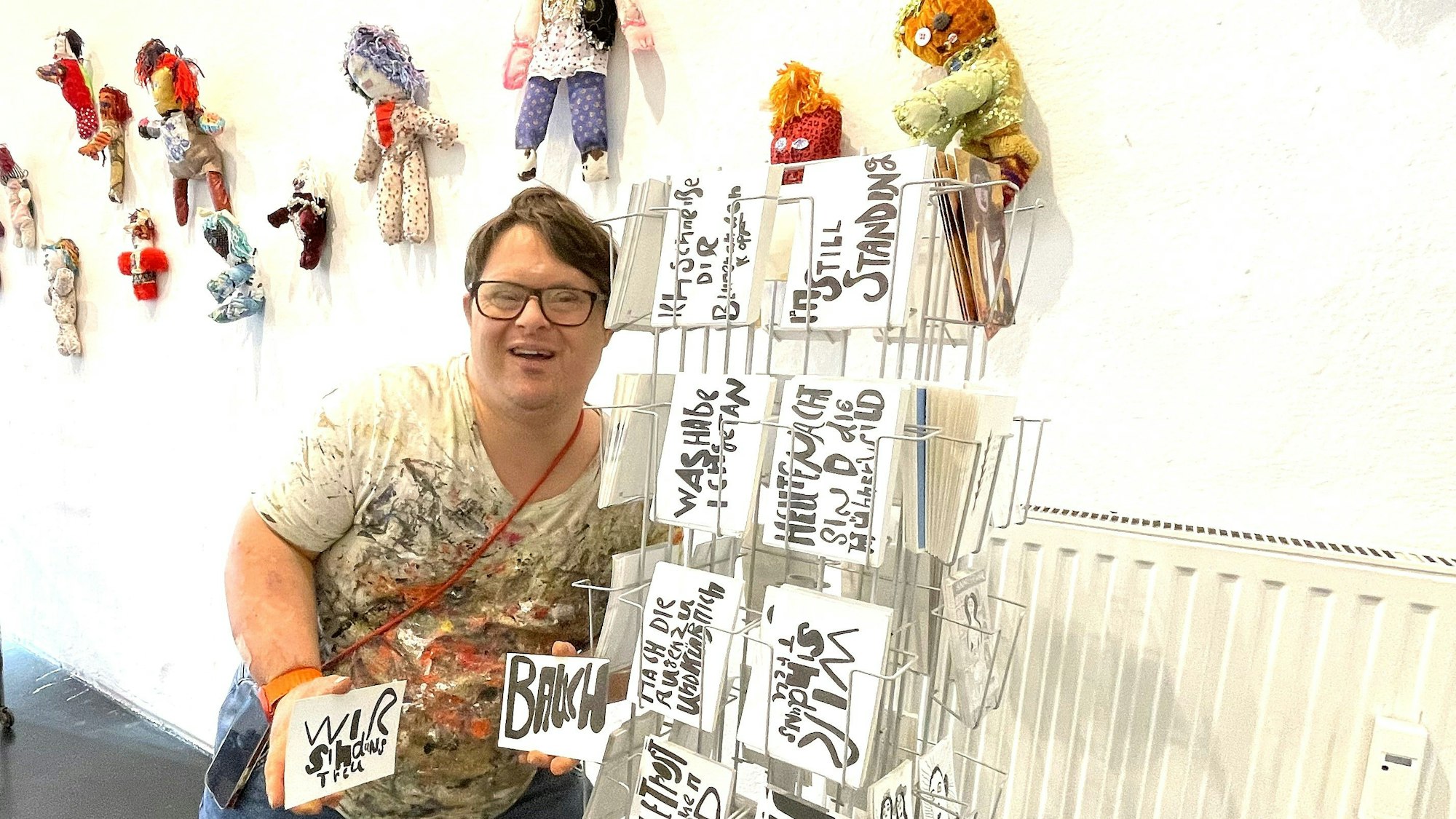 Ein junger Mann steht neben einem Postkartenständer. An der Wand hängen einzelne Püppchen.