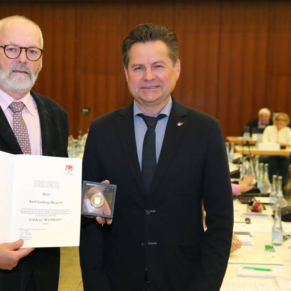 Zwei Männer, der linke hält eine Urkunde und eine Münze.