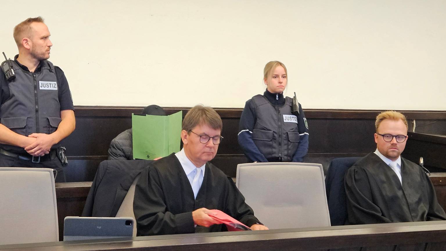 Zu sehen ist ein Mann auf einer Anklagebank, der einen Ordner vor sein Gesicht hält, außerdem sein Anwalt und Gerichtsvollzieher.