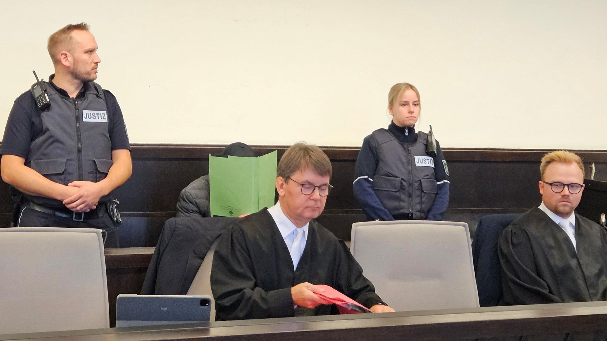 Zu sehen ist ein Mann auf einer Anklagebank, der einen Ordner vor sein Gesicht hält, außerdem sein Anwalt und Gerichtsvollzieher.