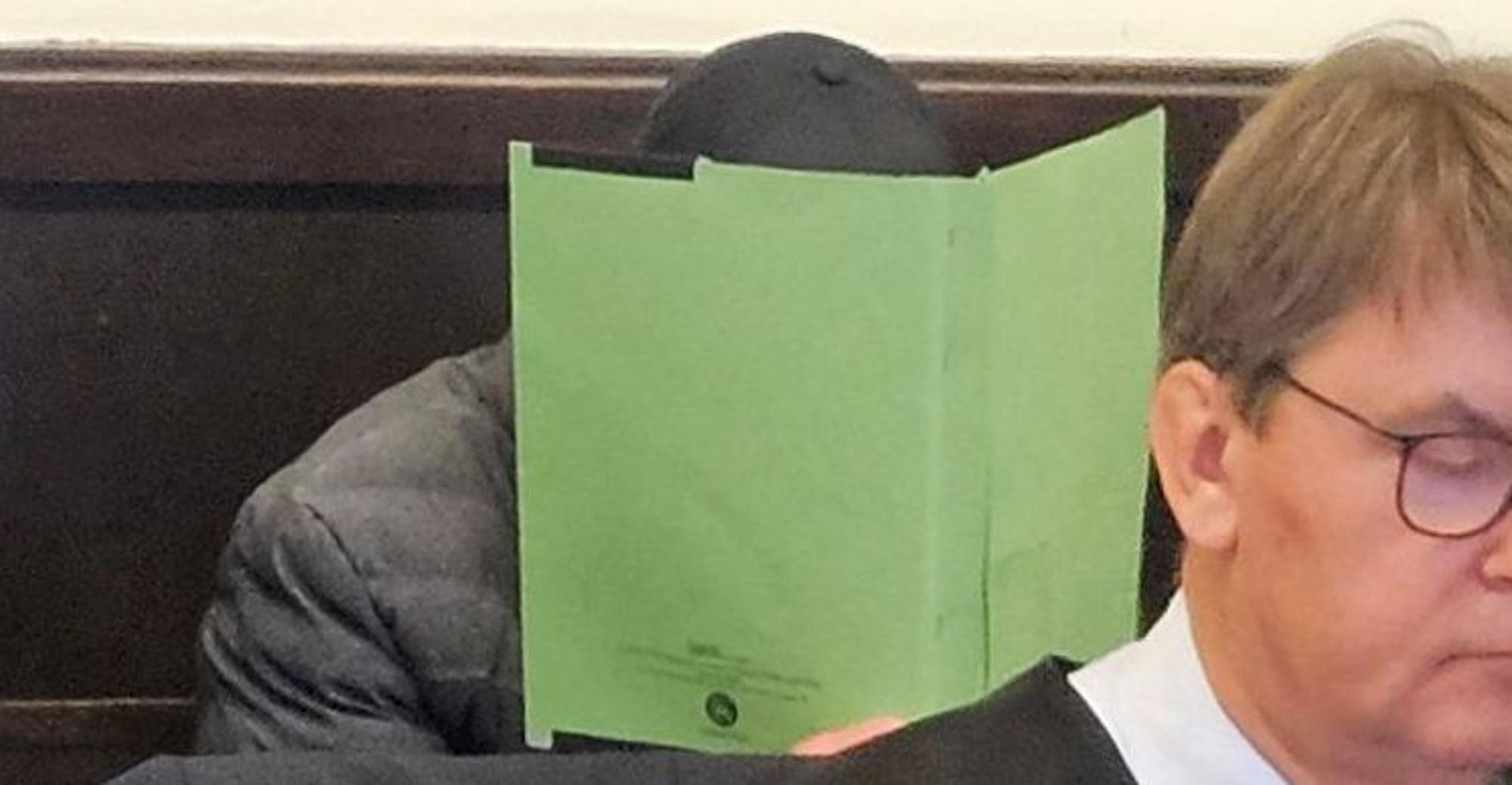Ein Angeklagter sitzt mit einem Aktendeckel vor dem Gesicht auf der Anklagebank des Paderborner Landgerichts.
