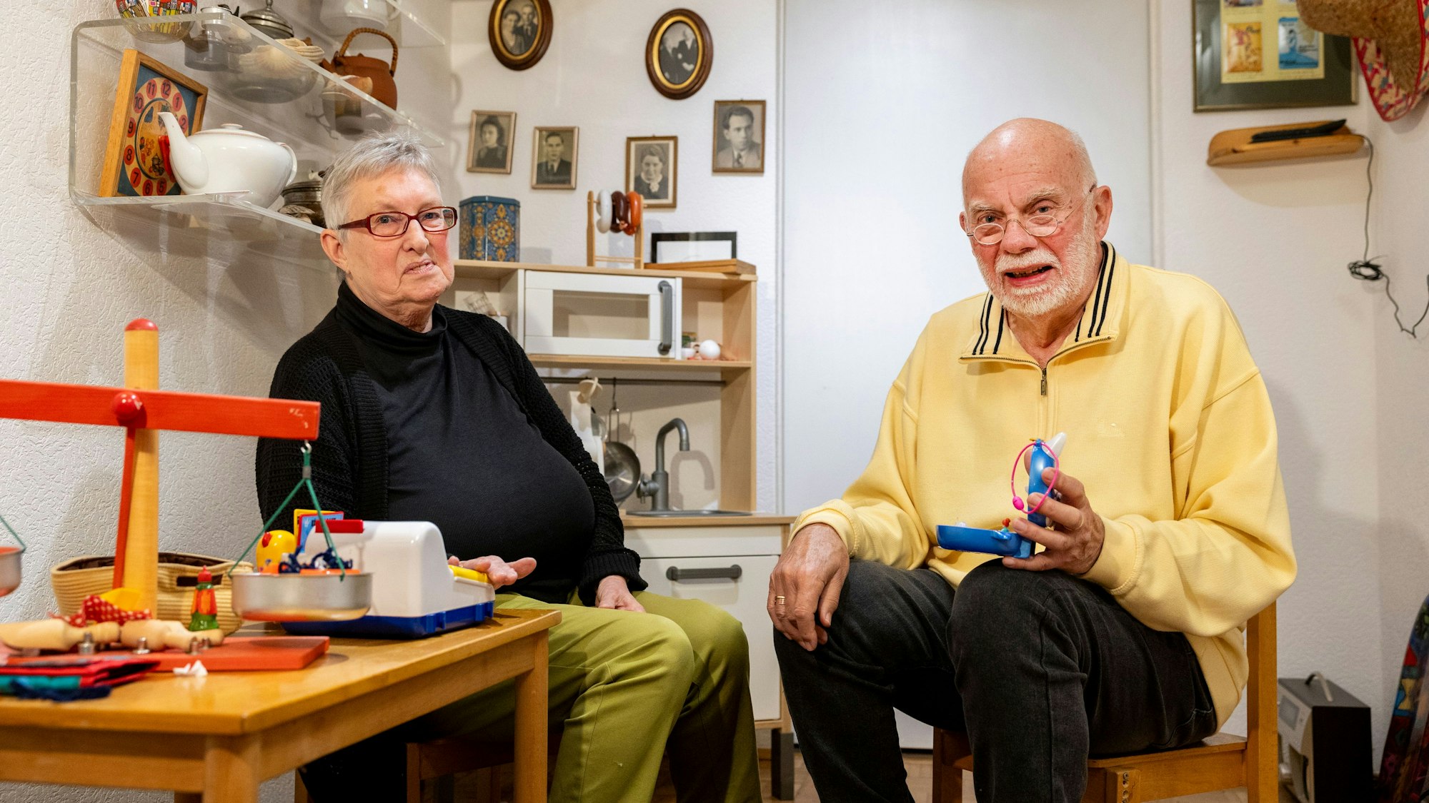 Gabriele und Werner Deuß sind inmitten von Spielsachen zu sehen. Sie sitzen auf Kinderstühlen.