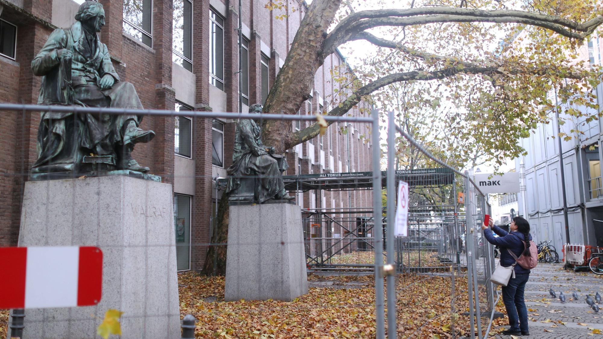 05.11.2025: Die hinter Bauzäunen „eingesperrten“ Statuen von Ferdinand Franz Wallraf (links) und Johann Heinrich Richartz vor dem Museum für Angewandte Kunst Köln. Hier stand der Vorgängerbau des nach beiden Mäzenen benannten Kunstmuseums.
