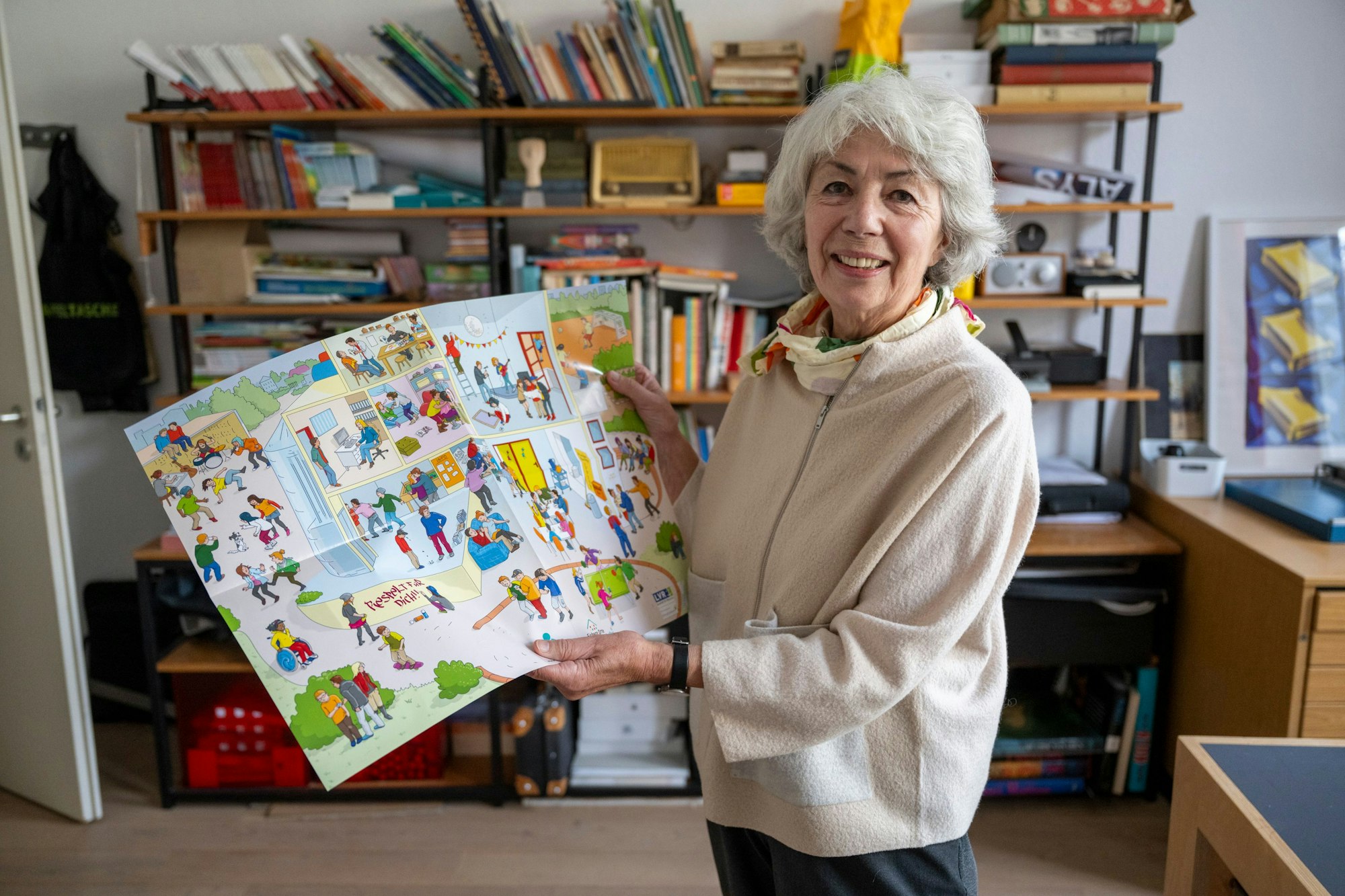 Die Kölner Illustratorin Dorothee Wolters steht vor einem Bücherregal und hält ein von ihr gestaltetes Wimmelbild-Plakat in den Händen.