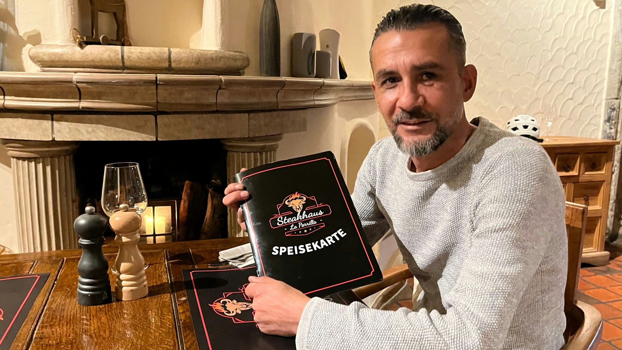 Ein Mann mit Bart sitzt an einem Tisch, er hält eine Speisekarte in die Kamera.