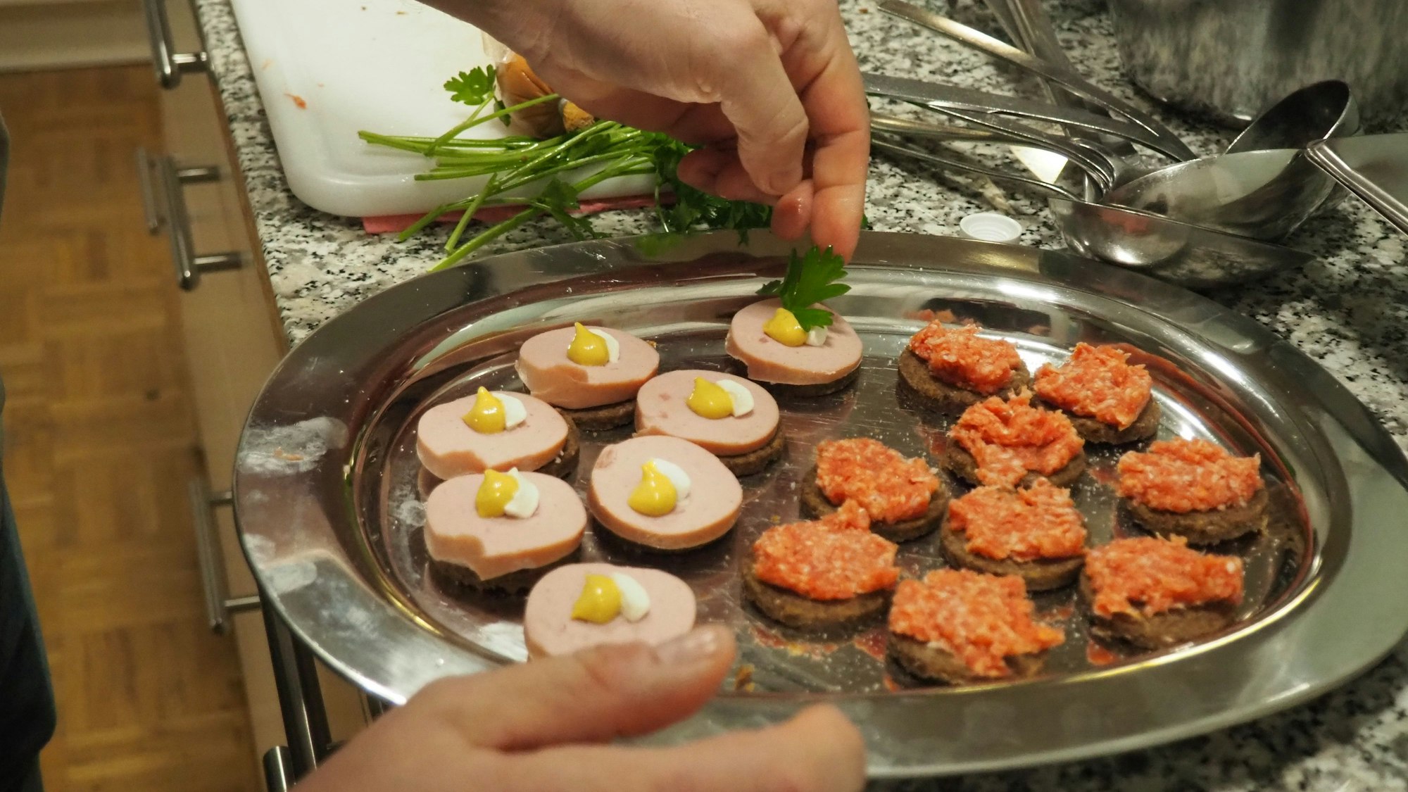 Das Bild zeigt eine Platte mit kleinen Häppchen, eine Hand fügt ein Petersilienblatt hinzu.
