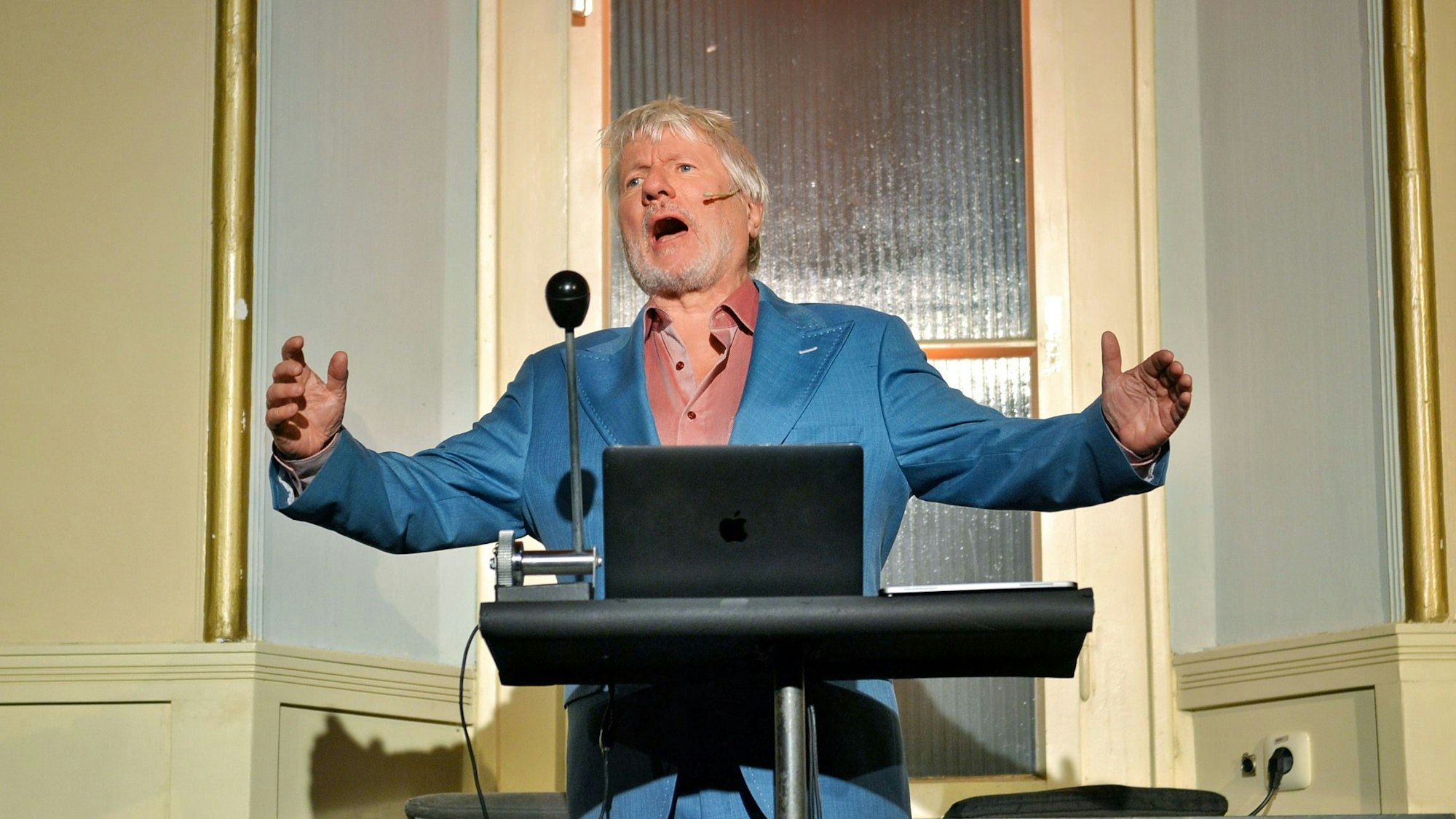 Jürgen Becker trägt einen blauen Anzug und hat Arme und Mund weit geöffnet.