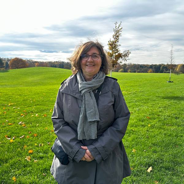 Ein Frau, die einen grauen Schal und Anorak trägt, steht auf einer weiten, hügeligen Wiese.