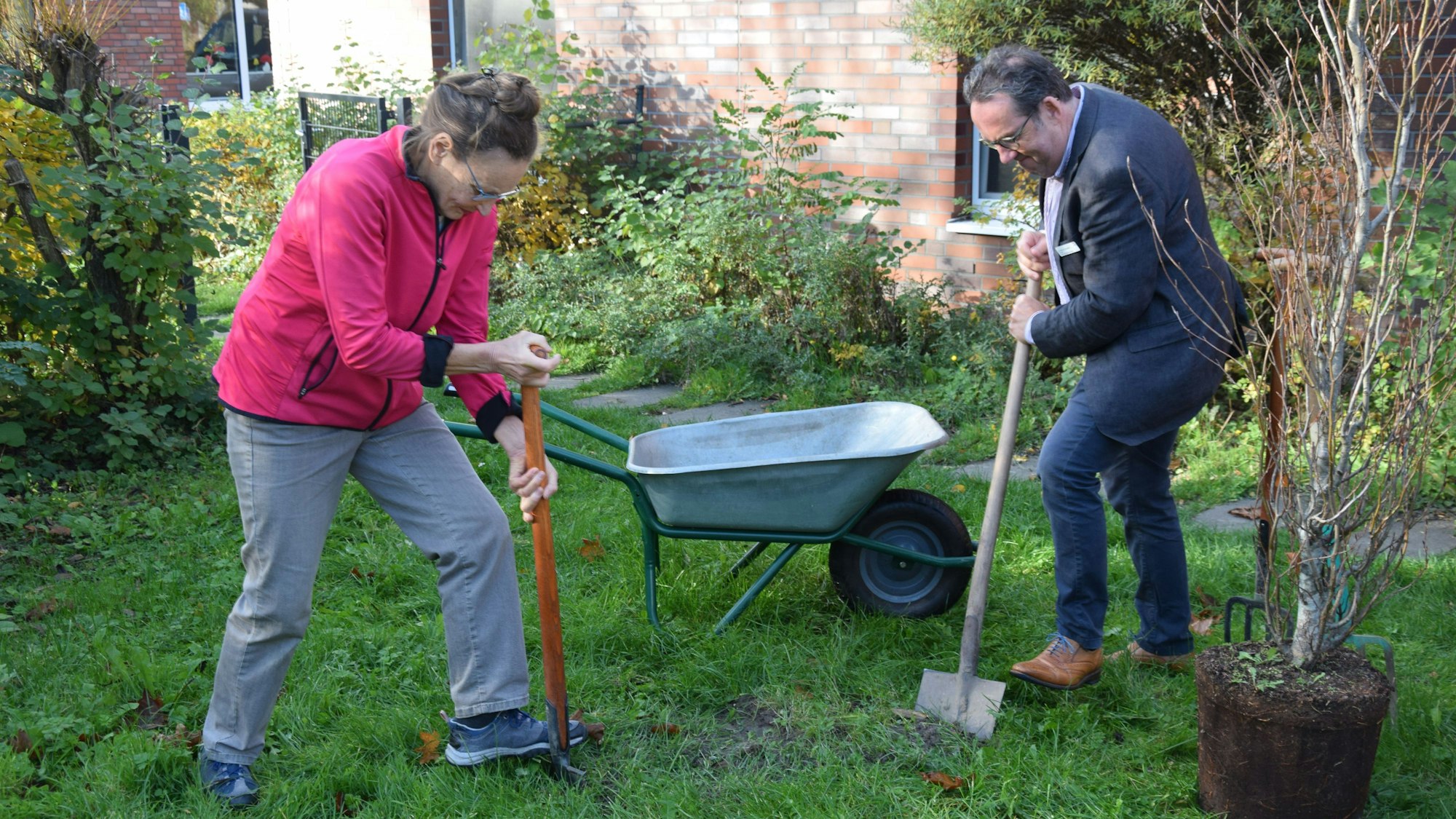 Eine Frau in pinker Jacke und grauer Hose und ein Mann im Anzug setzen den ersten Spatenstich für einen Baum.