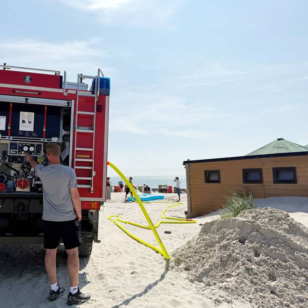 Ein Mann am Feuerwehrauto, ein Schlauch führt zum Strand.