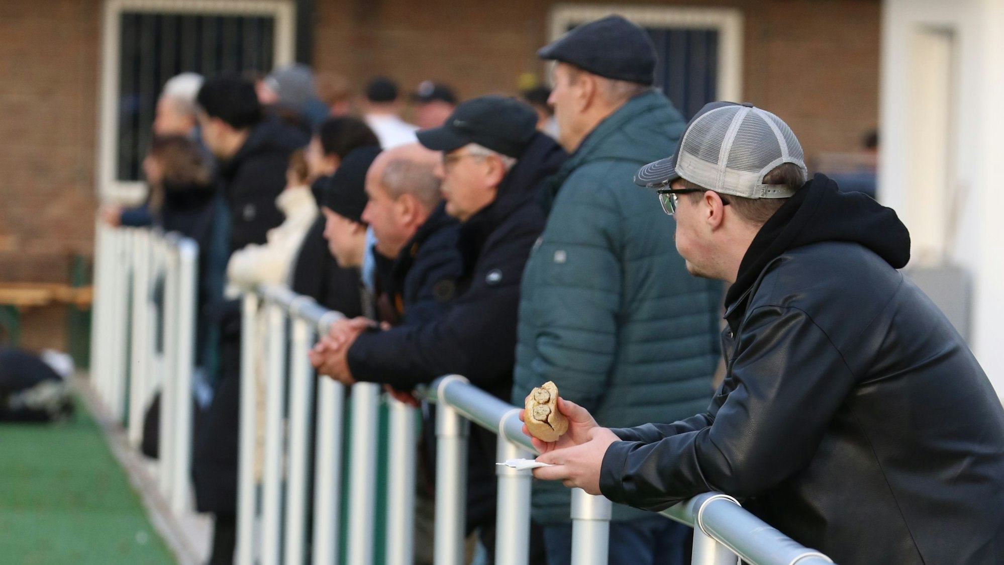 Zuschauer beobachten von der Reling das Spiel. Der vordere hält ein Bratwurstbrötchen in der Hand.