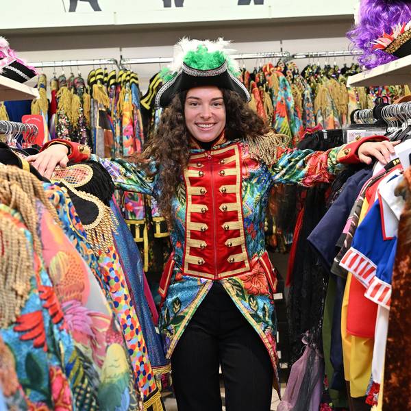 Eine Frau steht zwischen zwei Kleiderstangen an denen diverse Karnevalskostüme hängen. Sie trägt eine bunte Garde-Jacke und einen schwarzen Hut mit grünen Federn.