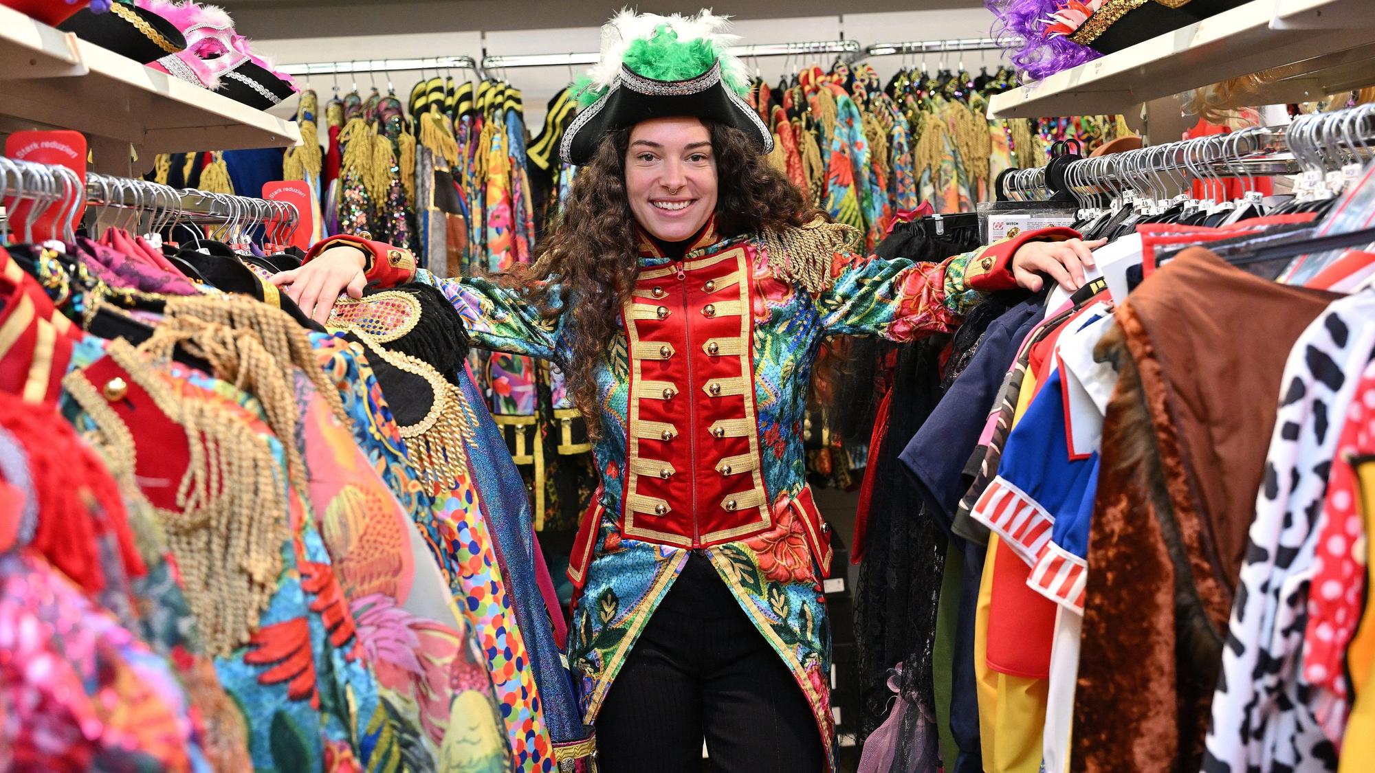 Eine Frau steht zwischen zwei Kleiderstangen an denen diverse Karnevalskostüme hängen. Sie trägt eine bunte Garde-Jacke und einen schwarzen Hut mit grünen Federn.
