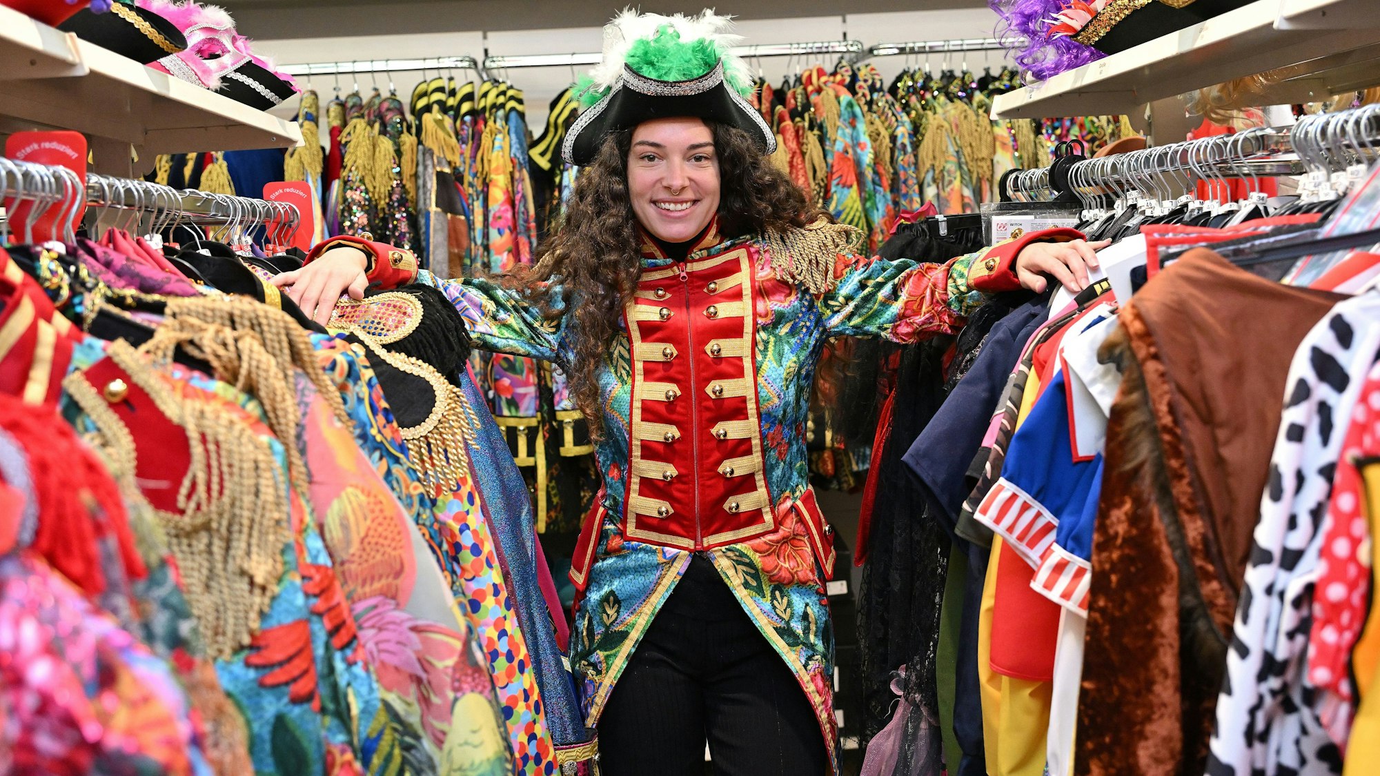 Eine Frau steht zwischen zwei Kleiderstangen an denen diverse Karnevalskostüme hängen. Sie trägt eine bunte Garde-Jacke und einen schwarzen Hut mit grünen Federn.