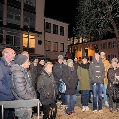 Am Mahnmal in Schleiden stehen Teilnehmende der Gedenkveranstaltung und hören F.A. Heinen bei seiner Rede zu.