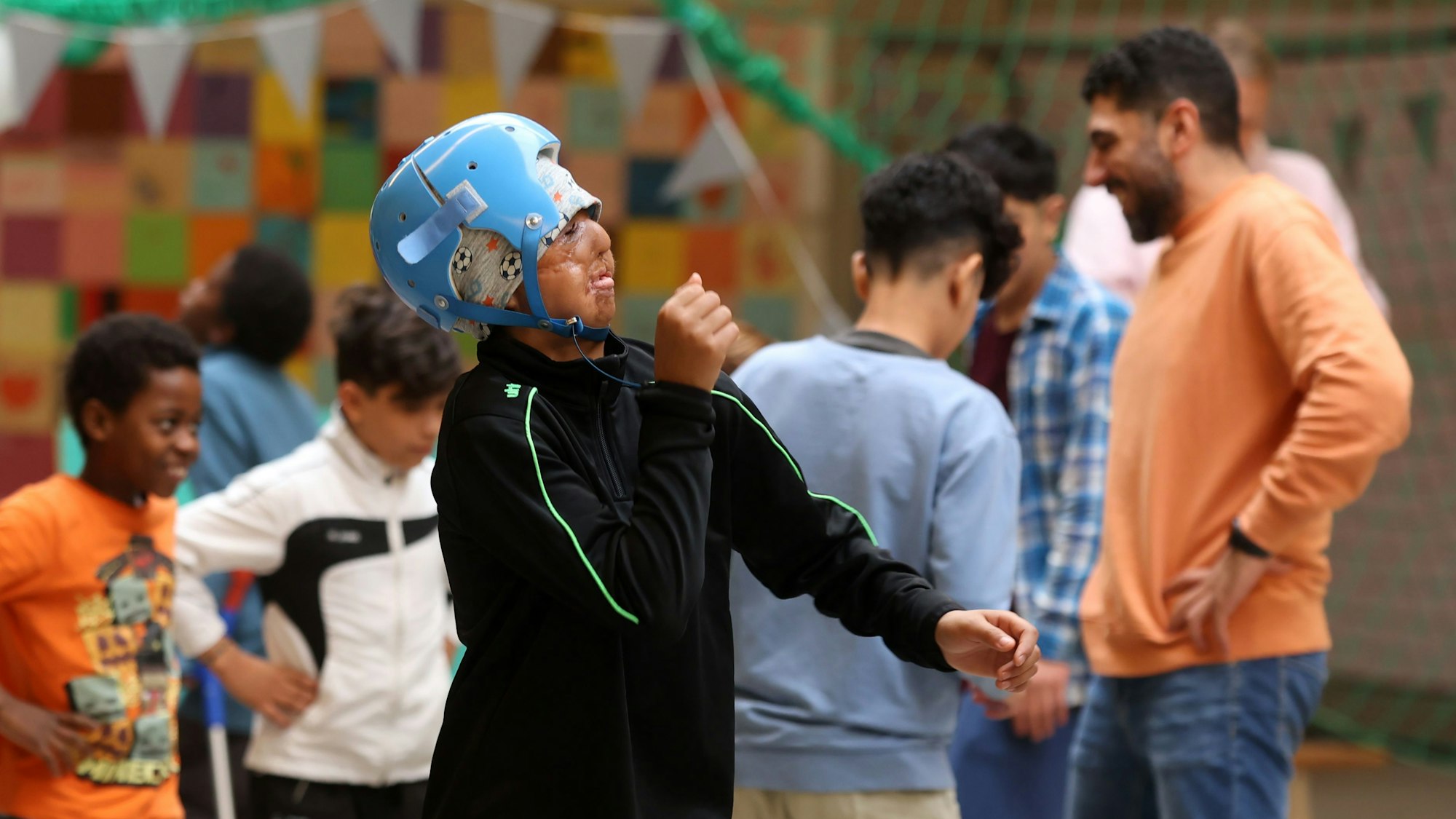 Abschiedfeier in der Turnhalle mit Kindern und Erwachsenen. Ein junge mit hellblauem Schutzhelm tanzt.