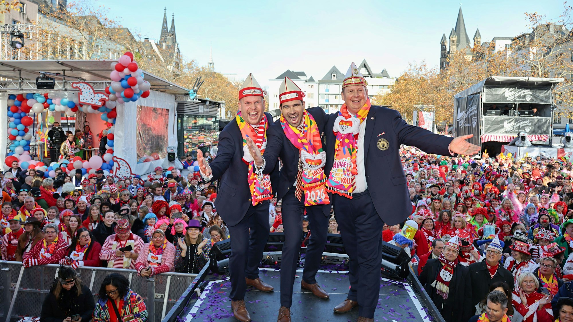 Drei Männer im Anzug und Karnevalsmütze und -schal stehen auf einer Bühne. Hinter ihnen eine große Menge an Menschen.