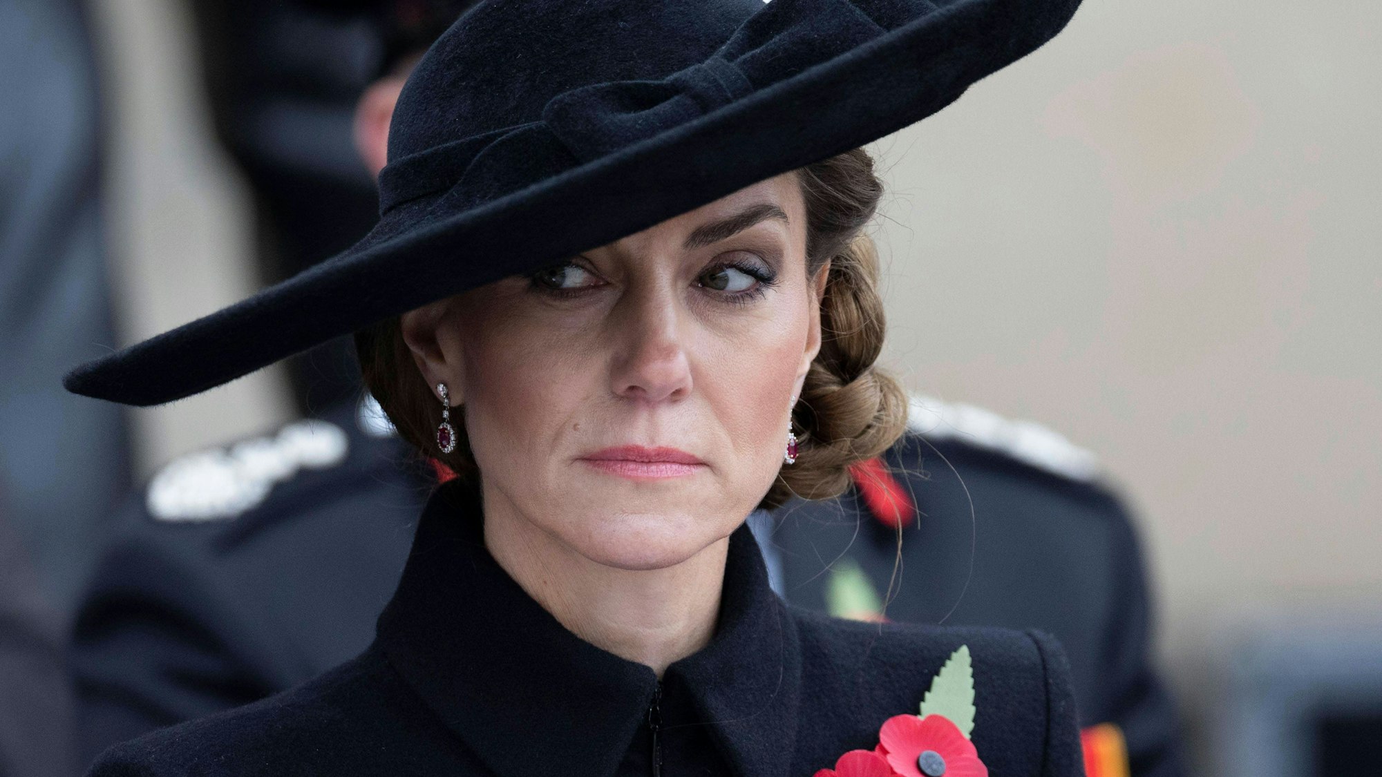 Prinzessin Kate mit ernstem Blick, schwarzem Hut und Mohnblumen am Mantel.