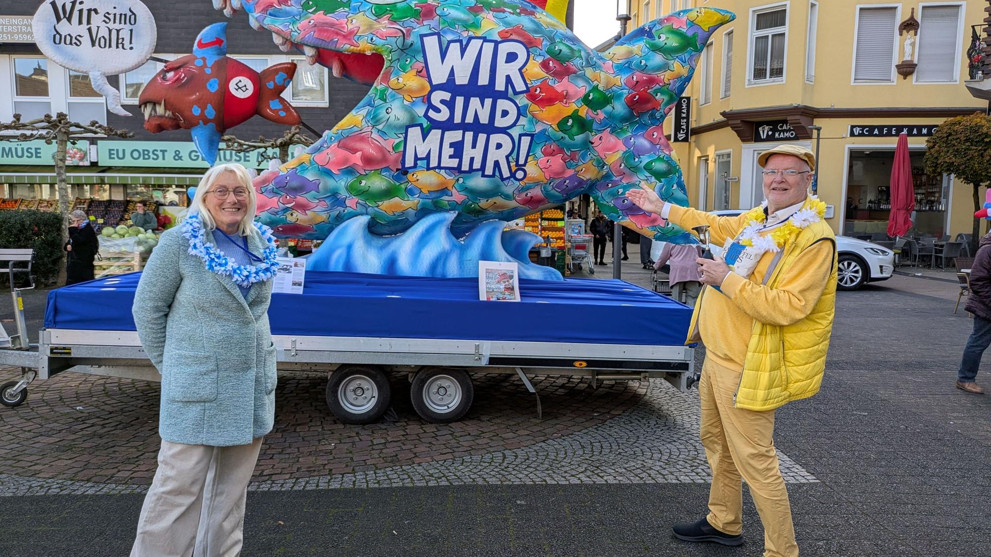 Andrea und Jo Johänning stehen vor dem Wagen, der einen großen Fisch mit der Aufschrift „Wir sind mehr“ darstellt, der such wiederum einen kleinen blau, rot und braunen Fisch schnappt.