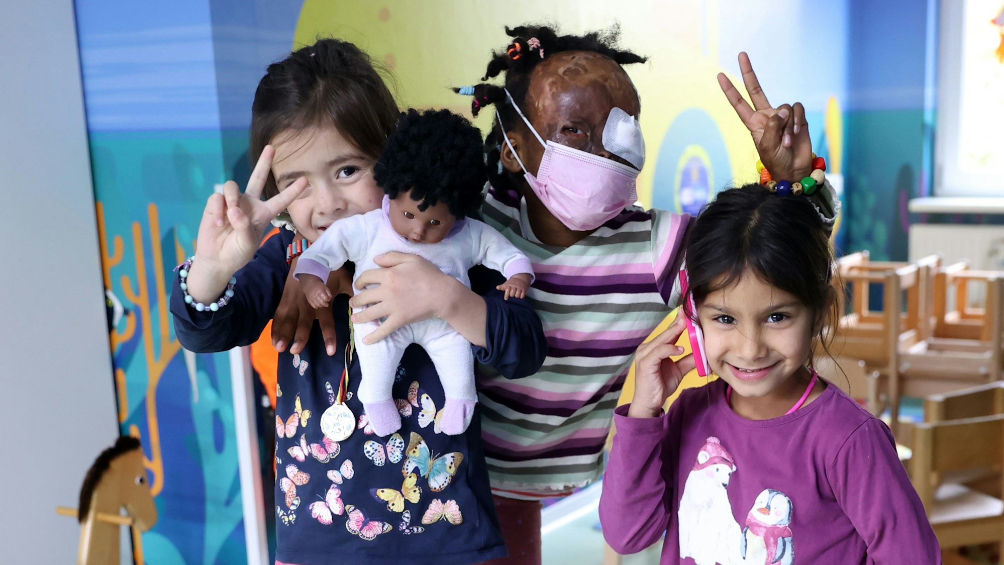 Drei Mädchen im Spielzimmer: zwei lachen offensichtlich in die Kamera, ein Mädchen tägt einen rosa Mundschutz und ein Augenpflaster.